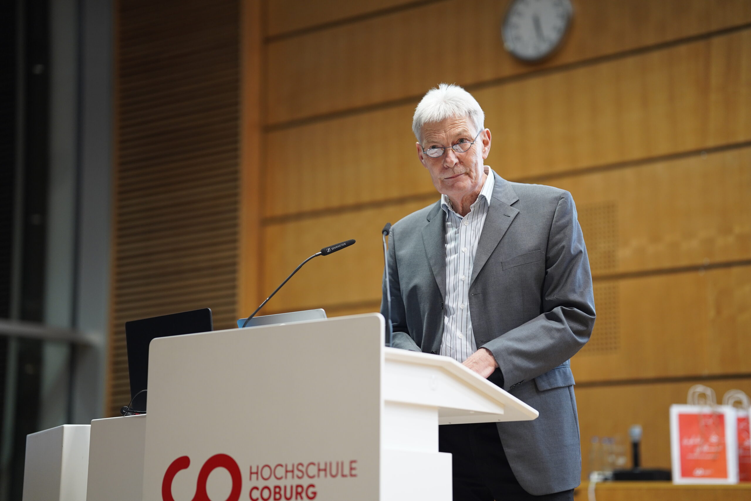 Ein älterer Mann in grauem Anzug und gestreiftem Hemd steht an einem Podium mit der Aufschrift "Hochschule Coburg" und spricht während des Dies academicus 2025 in einem Hörsaal mit Holzwänden und einer Uhr im Hintergrund. at Hochschule Coburg