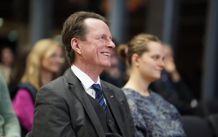 Ein Mann in Anzug und Krawatte lächelt, während er im Publikum des Dies academicus 2025 der Hochschule sitzt, während andere Personen im Hintergrund aufmerksam zusehen. at Hochschule Coburg