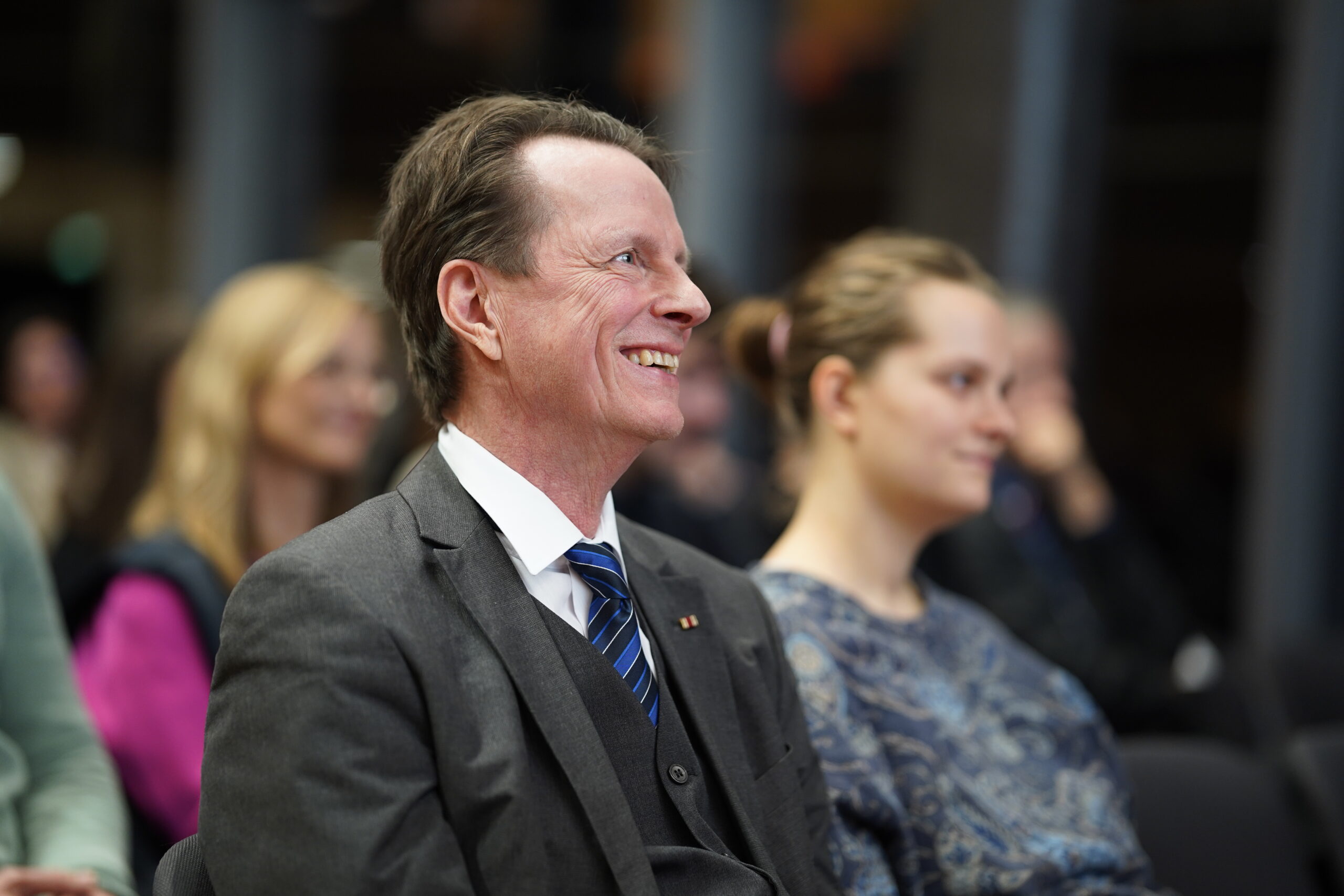 Ein Mann in Anzug und Krawatte lächelt, während er im Publikum des Dies academicus 2025 der Hochschule sitzt, während andere Personen im Hintergrund aufmerksam zusehen. at Hochschule Coburg