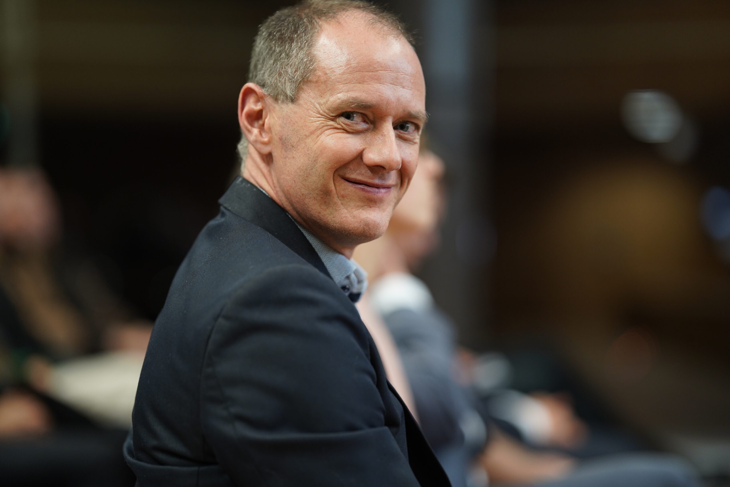 Ein Mann mittleren Alters in einem Anzug sitzt und lächelt leicht, während er in einer unscharfen Innenansicht einer Hochschule über die Schulter blickt, was die Atmosphäre des Dies academicus 2025 andeutet. at Hochschule Coburg