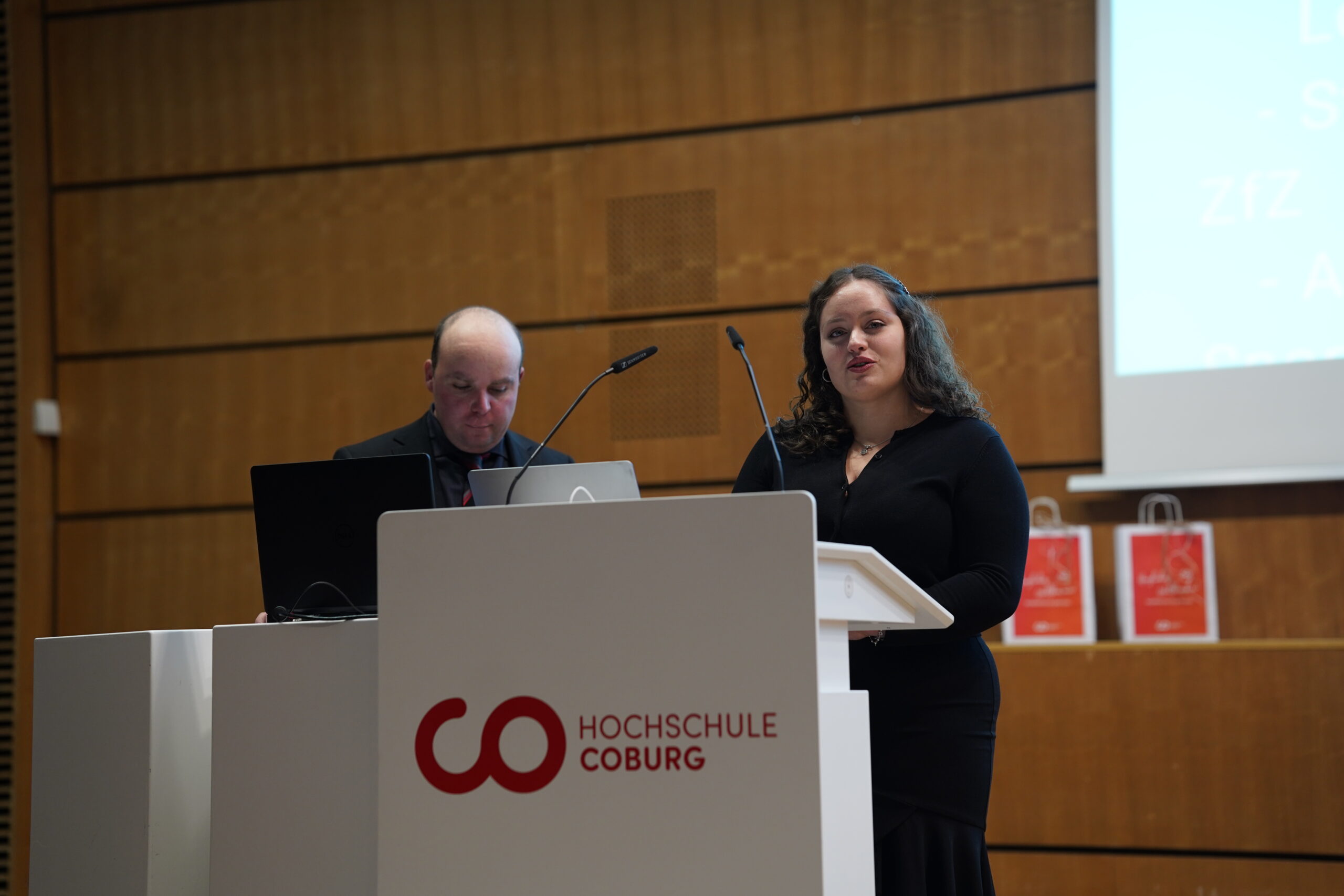 Eine Frau und ein Mann stehen an einem Podium mit der Aufschrift "Hochschule Coburg" in einem modernen Hörsaal während des Dies academicus 2025. Die Frau spricht, während der Mann auf einen Laptop schaut. Hinter ihnen stehen Holzwände und Präsentationsbildschirme. at Hochschule Coburg
