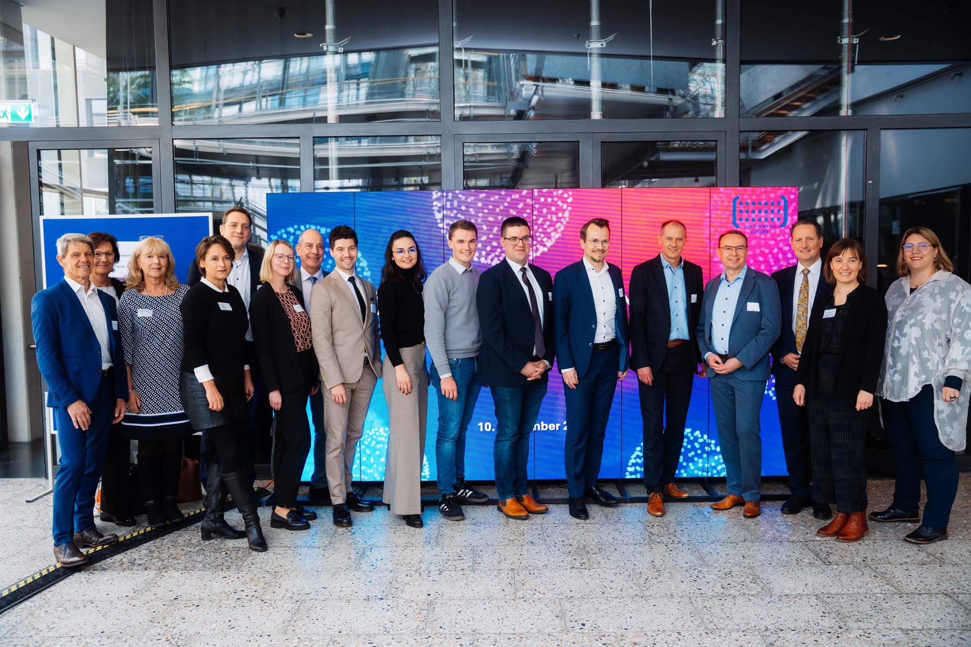 Eine Gruppe von fünfzehn professionell gekleideten Absolventen steht in einer Reihe und lächelt in die Kamera vor einer farbenfrohen digitalen Anzeige und Glastüren, die bayerische Hochschulen in einer modernen Innenumgebung repräsentieren. at Hochschule Coburg