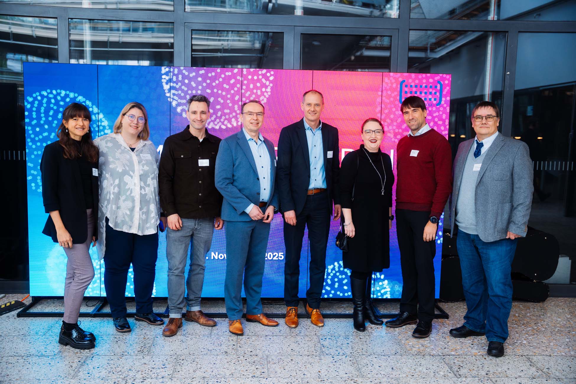 Acht Personen stehen in einer Reihe und lächeln in die Kamera. Sie befinden sich in einem Innenraum, sind leger gekleidet und haben einen farbenfrohen digitalen Bildschirm mit abstrakten Mustern im Hintergrund, der Absolventen bayerischer Hochschulen repräsentiert. at Hochschule Coburg