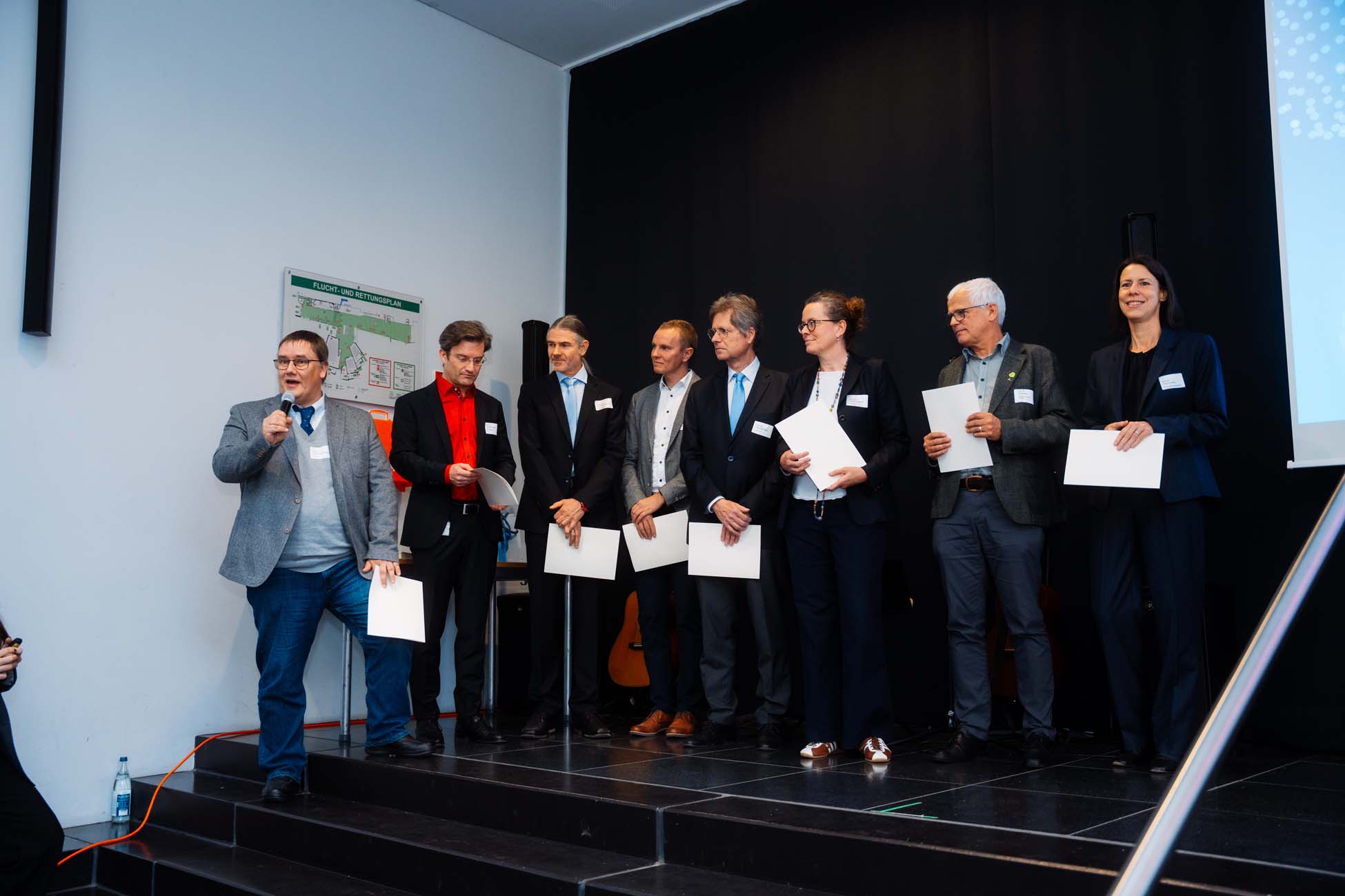 Eine Gruppe von acht Absolventen in formeller Kleidung steht auf einer Bühne und hält Urkunden in der Hand. Eine Person spricht in ein Mikrofon, während die anderen lächelnd ins Publikum schauen. Im Hintergrund sind ein schwarzer Vorhang und die Veranstaltungsbeschilderung der bayerischen Hochschulen zu sehen. at Hochschule Coburg