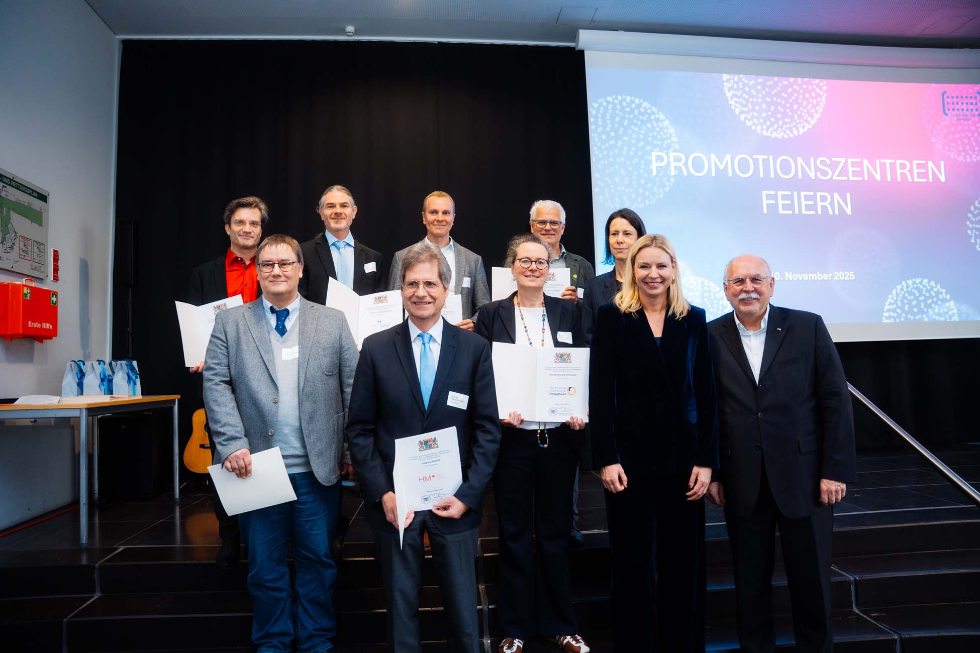 Eine Gruppe von Personen in formeller Kleidung posiert auf der Bühne, einige halten Urkunden in der Hand und feiern Absolventen der bayerischen Hochschulen. Eine Präsentationsleinwand hinter ihnen zeigt "PROMOTIONSZENTREN FEIERN" an, was eine Preisverleihung oder Anerkennungsveranstaltung zu sein scheint. at Hochschule Coburg