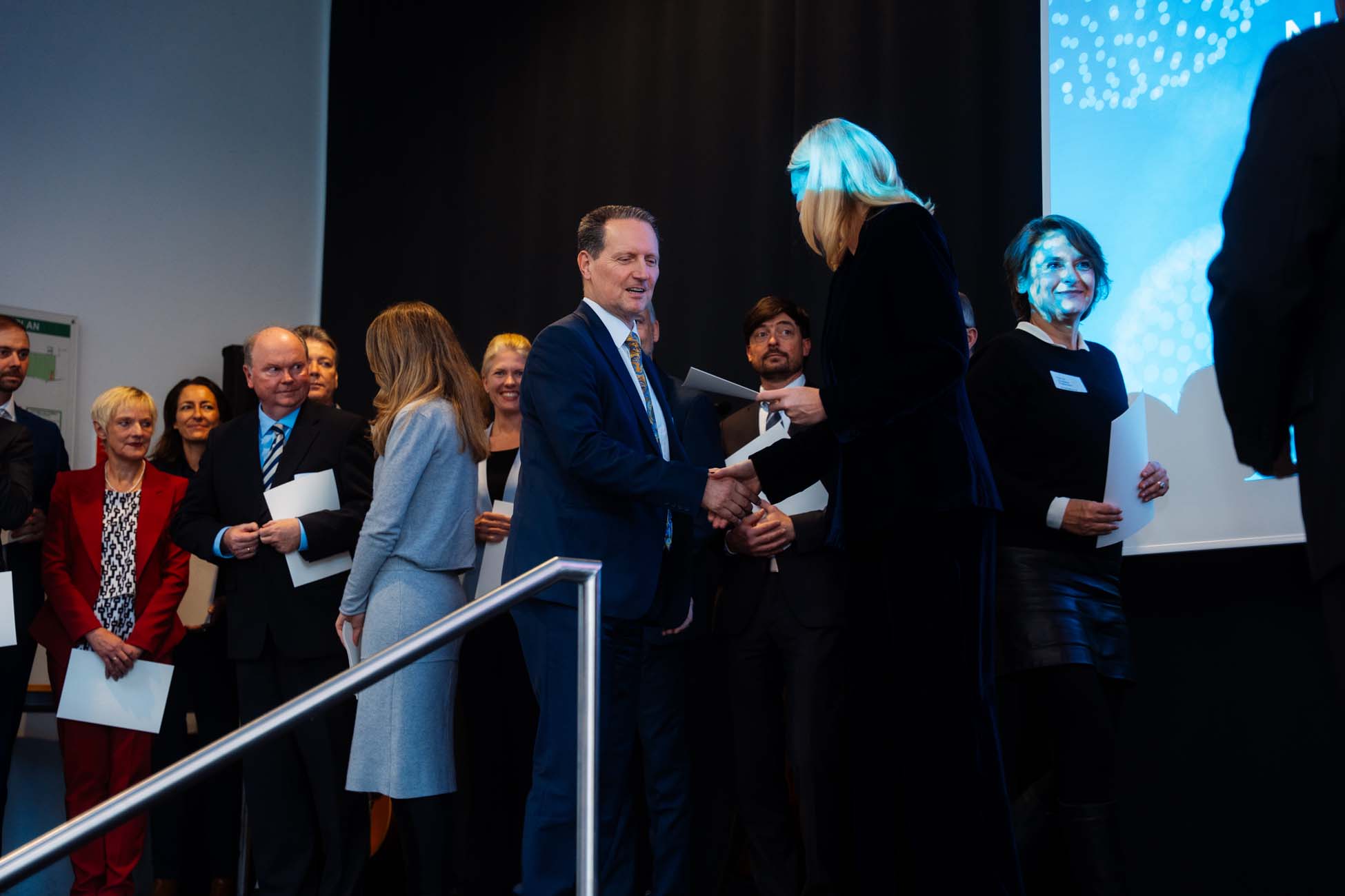 Eine Gruppe von Absolventen steht in einer Reihe auf der Bühne, einige halten Zertifikate in der Hand. Ein Mann im Anzug schüttelt einer Frau in Schwarz die Hand, während andere warten und zusehen. Vor einer Kulisse aus Vorhang und Leinwand werden die Leistungen an den bayerischen Hochschulen gewürdigt. at Hochschule Coburg