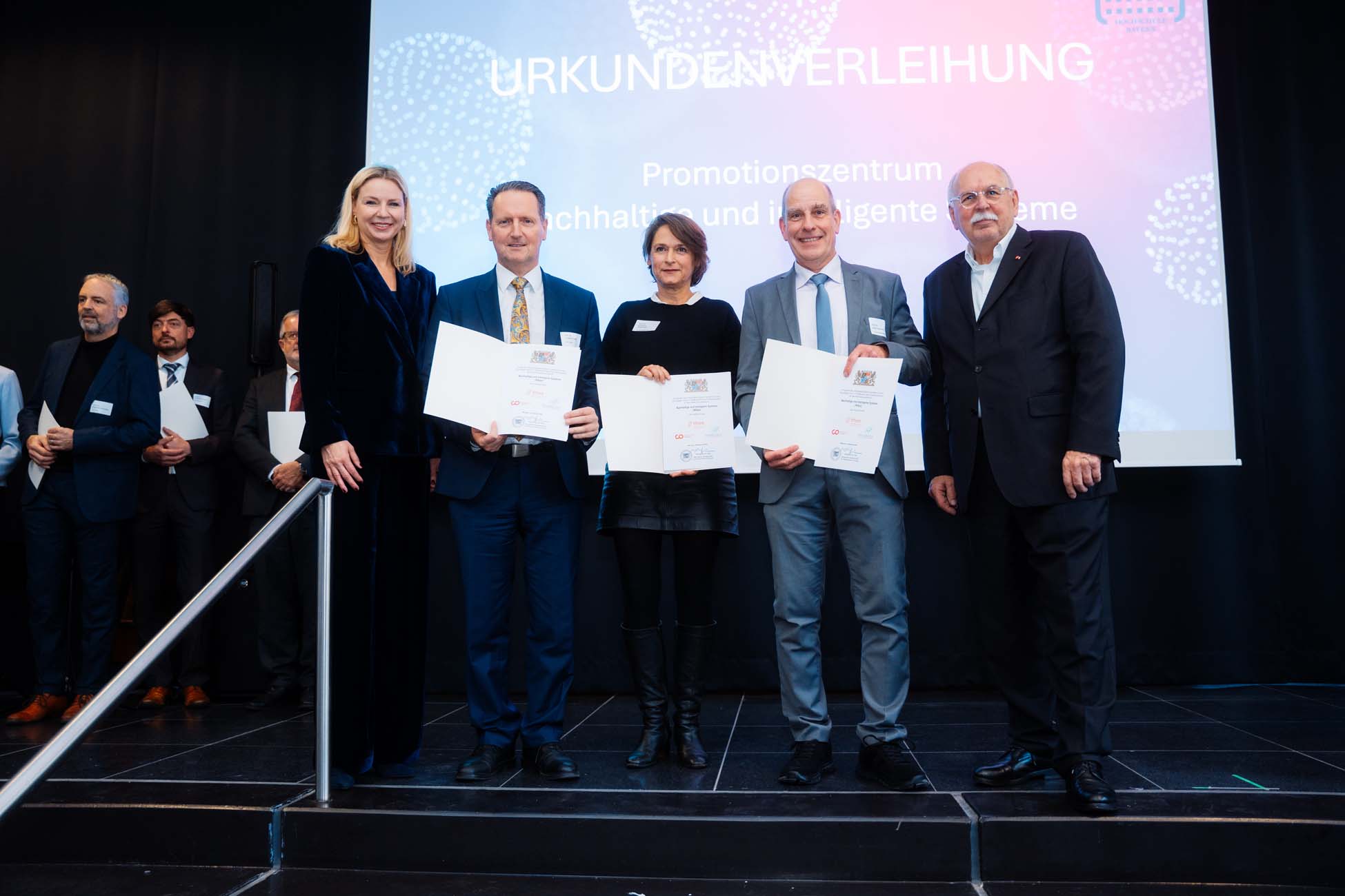 Fünf Absolventen stehen in förmlicher Kleidung auf der Bühne und halten Urkunden in den Händen, während andere zuschauen. Auf dem Präsentationsbildschirm im Hintergrund ist "Urkundenverleihung" und ein deutscher Text mit Bezug zu den bayerischen Hochschulen zu sehen. at Hochschule Coburg