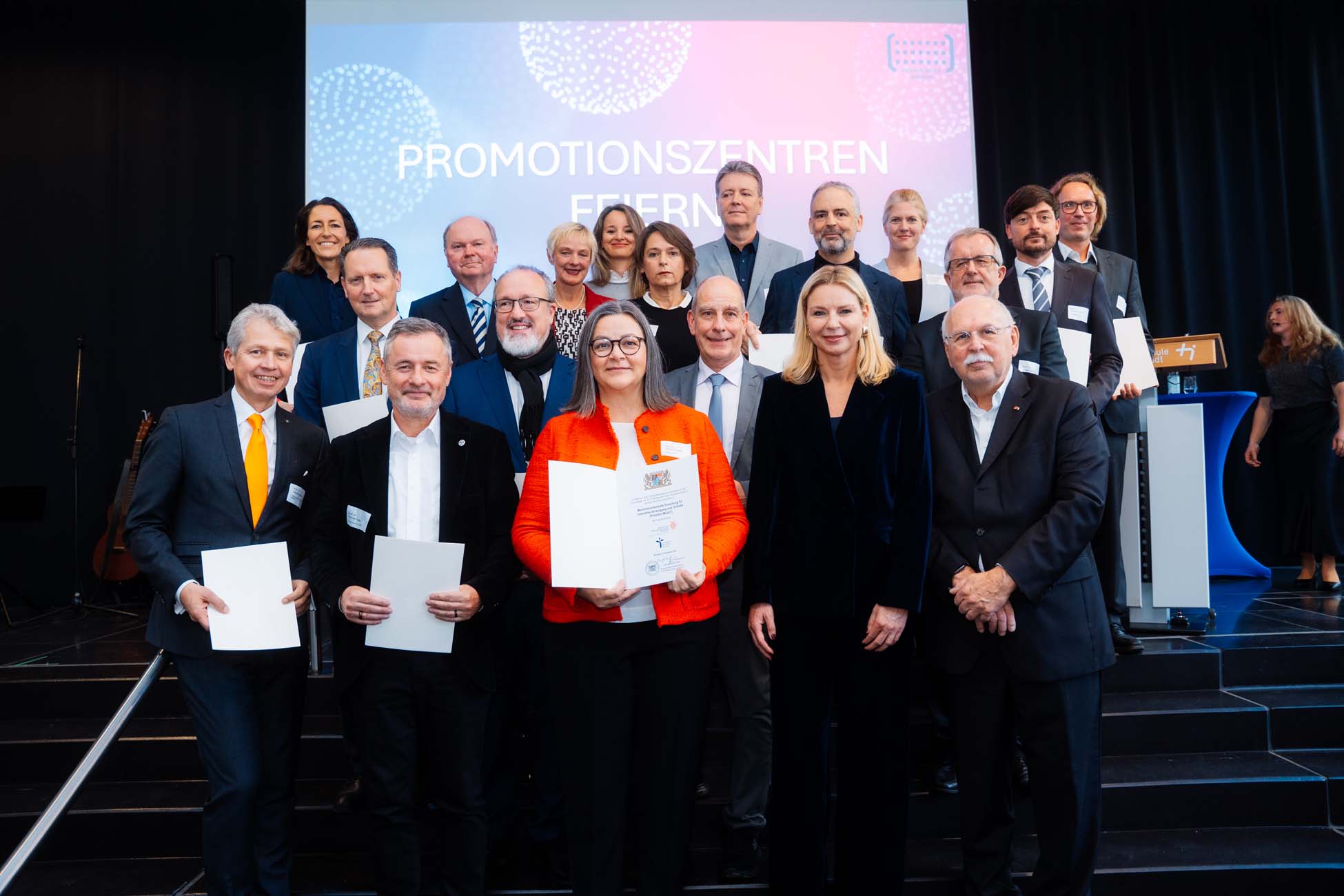 Eine Gruppe professionell gekleideter Absolventen steht auf der Bühne, lächelt und hält Urkunden in der Hand. Hinter ihnen prangt in großen Lettern das Wort "PROMOTIONSZENTREN", das die Leistungen der bayerischen Hochschulen hervorhebt. at Hochschule Coburg