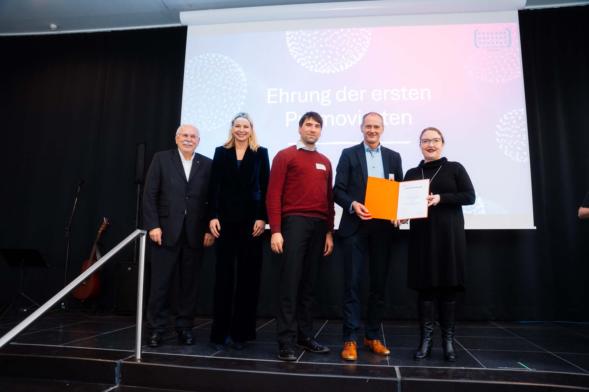 Fünf Personen stehen auf der Bühne, zwei halten eine orangefarbene Urkunde. Auf der Leinwand dahinter steht "Ehrung der ersten Promovierten". Sie alle lächeln, sind förmlich gekleidet und feiern die Absolventen der bayerischen Hochschulen. at Hochschule Coburg