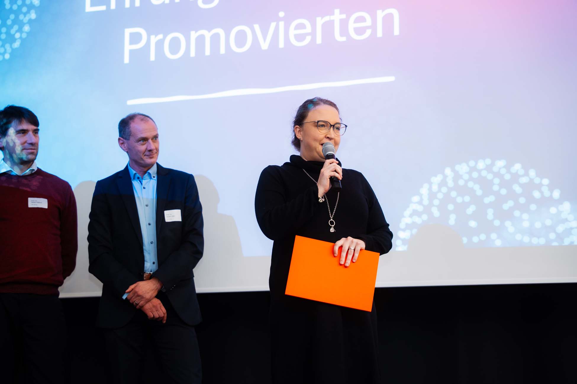 Eine Frau mit einem Mikrofon und einem orangefarbenen Ordner spricht auf der Bühne neben zwei Männern, während im Hintergrund eine Präsentationsfolie mit deutschem Text über bayerische Hochschulen projiziert wird. at Hochschule Coburg