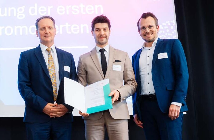 Fünf Absolventen in formeller Kleidung stehen während einer Preisverleihung auf der Bühne. Eine Person hält eine Urkunde in der Hand. Hinter ihnen ist auf einer Präsentationsfolie "Ehrung der ersten Promovierten" der bayerischen Hochschulen Promotionszentren zu sehen. at Hochschule Coburg