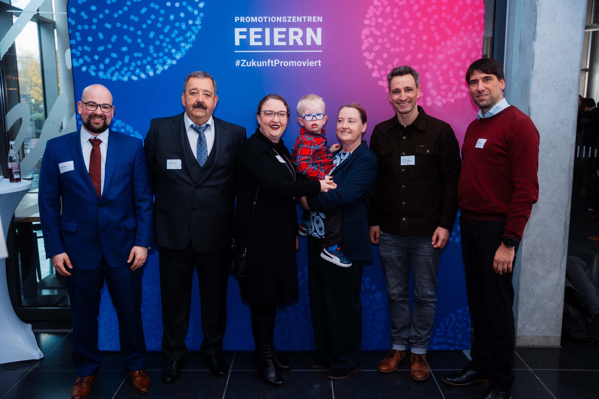 Eine Gruppe von sechs Erwachsenen und einem Kind in formeller und halbformeller Kleidung steht lächelnd vor einem farbenfrohen Hintergrund mit der Aufschrift "PROMOTIONSZENTREN FEIERN" und feiert Absolventen der bayerischen Hochschulen auf der Veranstaltung. at Hochschule Coburg