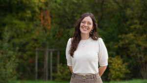 A woman with long brown hair, glasses, and a white sweater stands smiling outdoors in front of green trees, with her hands behind her back. at Hochschule Coburg