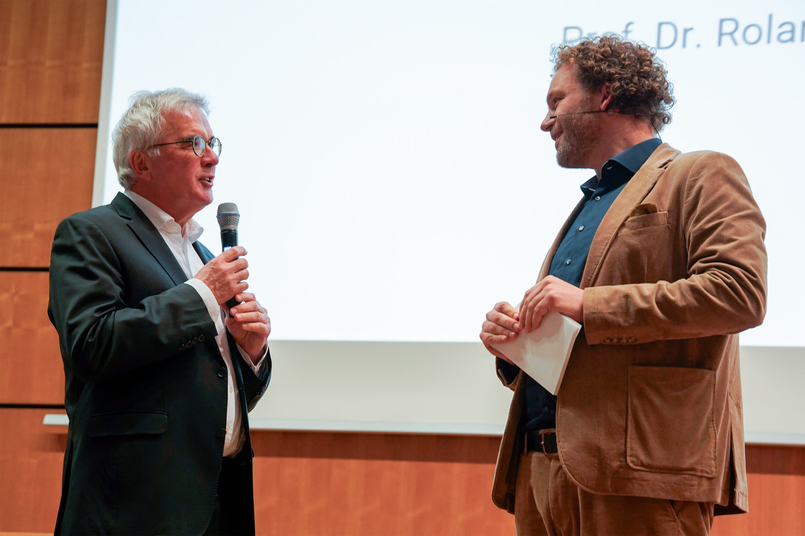 Bei der Veranstaltung Dies academicus 2025 der Hochschule stehen zwei Männer in Anzügen auf einer Bühne - einer spricht in ein Mikrofon, der andere hört aufmerksam zu und hält Papiere in der Hand, beide stehen vor einer großen Projektionsfläche, auf der ein Teil des Textes zu sehen ist. at Hochschule Coburg