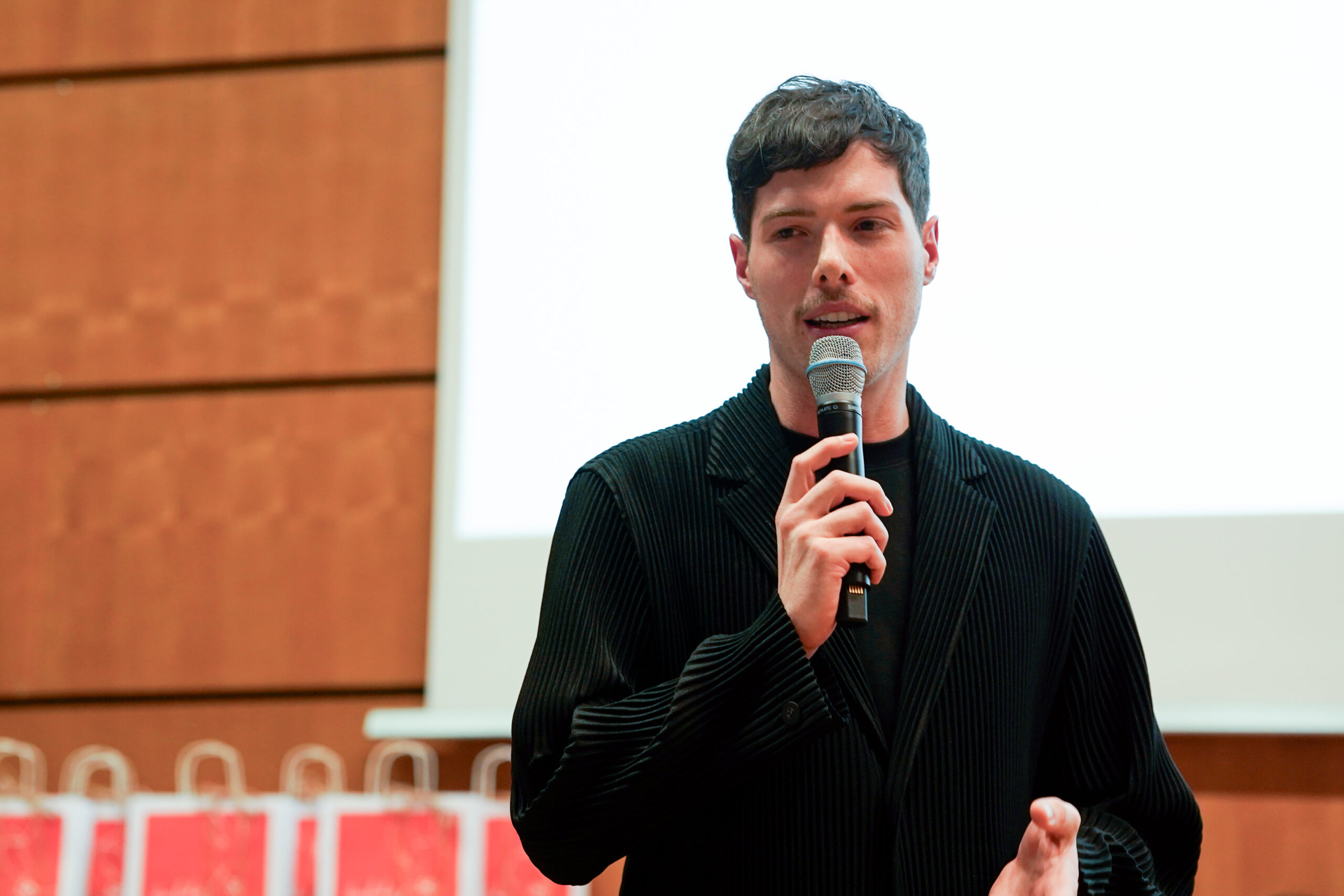 Ein Mann mit kurzen dunklen Haaren, der eine schwarze Jacke trägt, spricht in ein Mikrofon, während er vor einer Projektionsfläche am Dies academicus 2025 der Hochschule steht. Im Hintergrund sind Geschenktüten zu sehen. at Hochschule Coburg