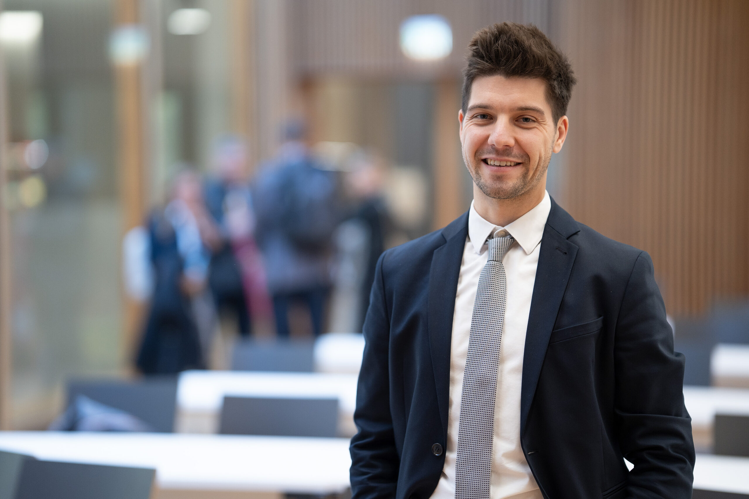 Ein junger Mann in Anzug und Krawatte steht lächelnd in einem modernen, hell erleuchteten Büro und verkörpert die Zukunft der Wirtschaft, während im Hintergrund verschwommene Kollegen über Elektromobilität diskutieren. at Hochschule Coburg