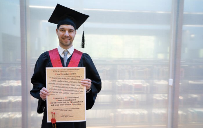 Ein junger Mann mit Kappe und Talar lächelt, während er vor einem Bücherregal ein Abschlusszeugnis in der Hand hält. Das deutet auf eine Bibliothek und eine strahlende Zukunft hin, vielleicht in Bereichen wie Elektromobilität oder Stromnetzinnovation. at Hochschule Coburg