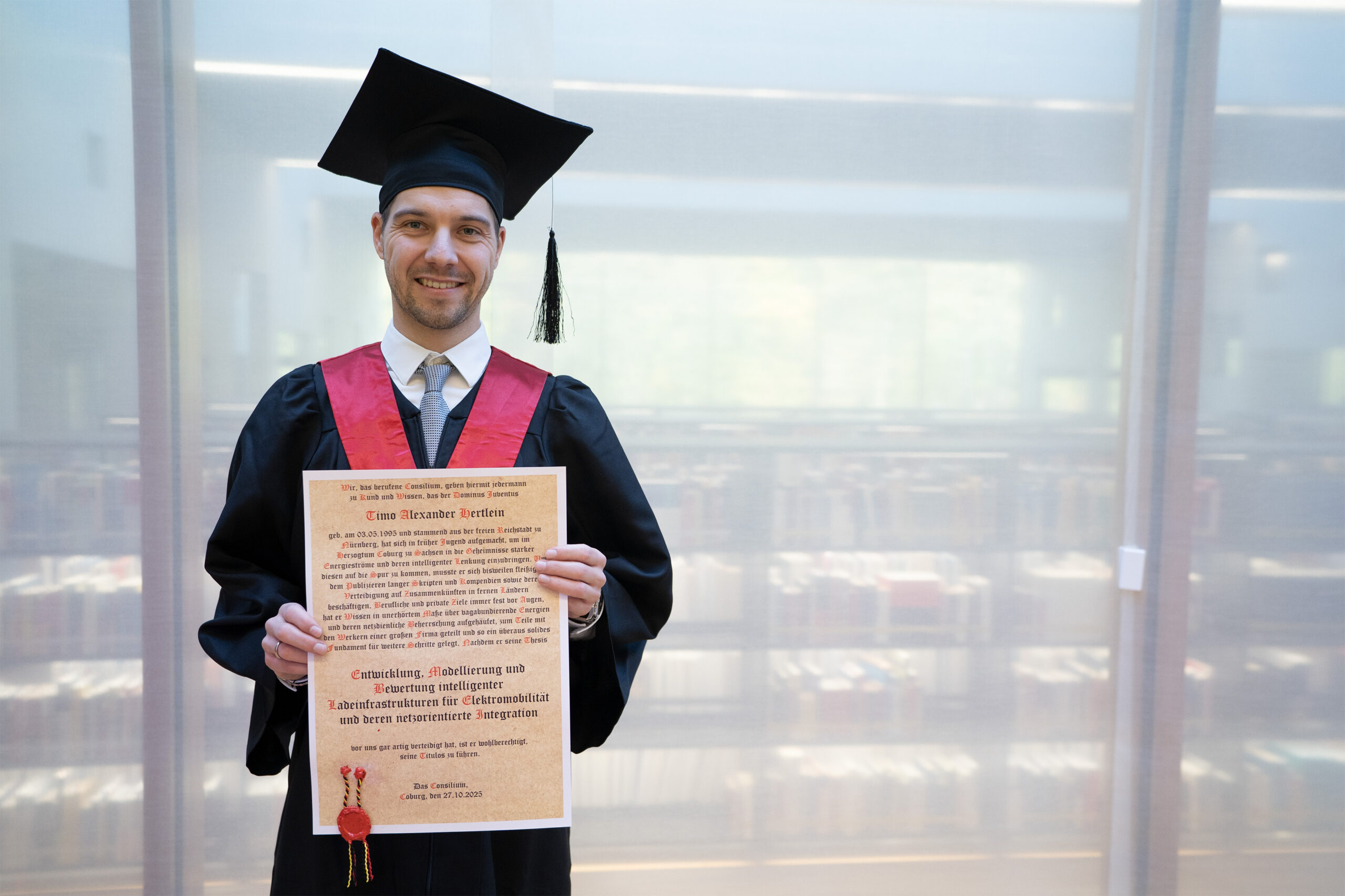 Ein junger Mann mit Kappe und Talar lächelt, während er vor einem Bücherregal ein Abschlusszeugnis in der Hand hält. Das deutet auf eine Bibliothek und eine strahlende Zukunft hin, vielleicht in Bereichen wie Elektromobilität oder Stromnetzinnovation. at Hochschule Coburg
