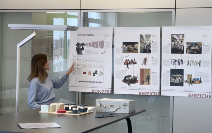 Eine Frau steht neben einem Tisch und zeigt auf ein Plakat über "Bürokonzepte der Zukunft" mit Diagrammen und Bildern. Zwei weitere Plakate, eines über den CREAPOLIS-Preis, sind ebenfalls zu sehen. Auf dem Tisch liegen verschiedene Materialien und ein weißer Koffer. at Hochschule Coburg