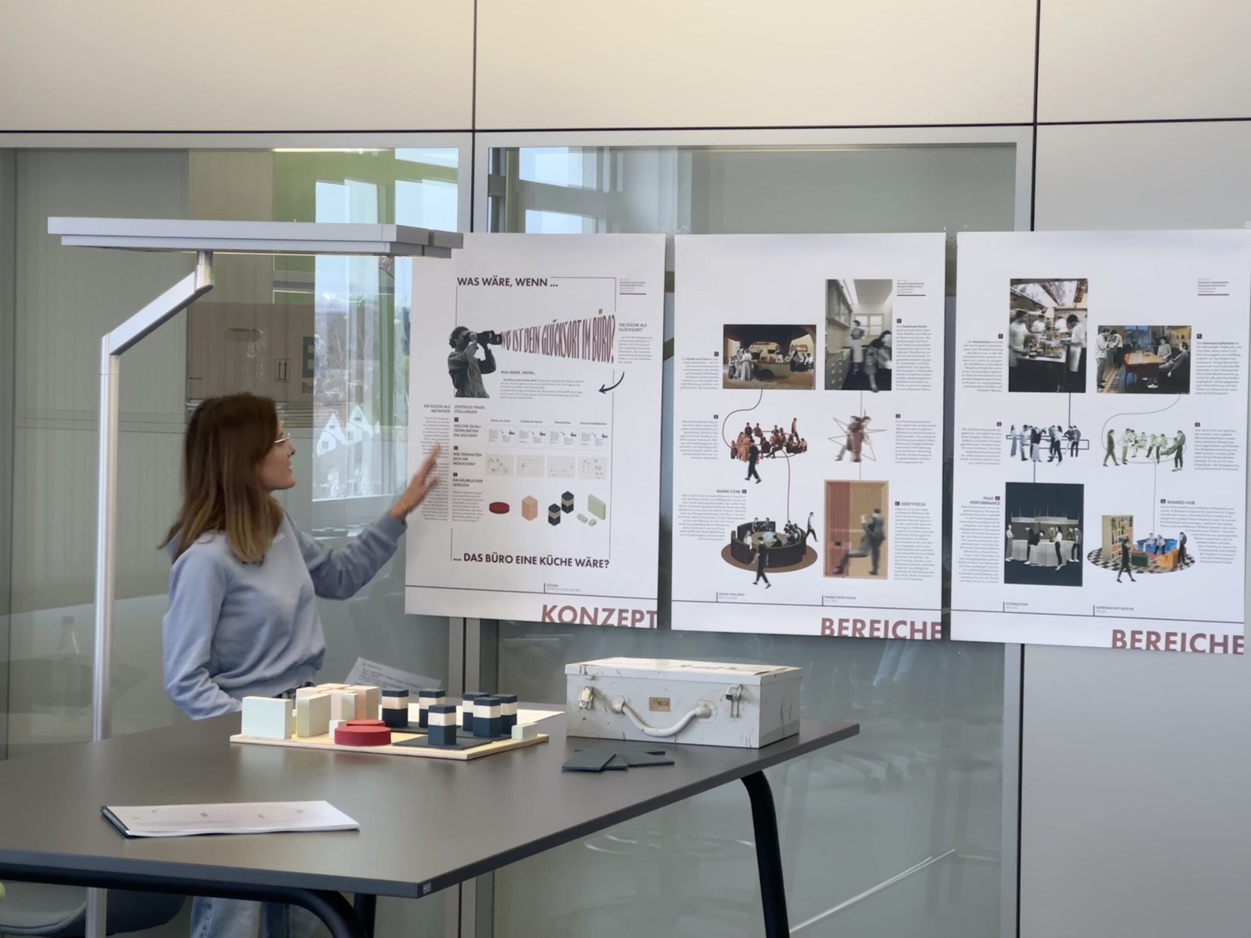 Eine Frau steht neben einem Tisch und zeigt auf ein Plakat über "Bürokonzepte der Zukunft" mit Diagrammen und Bildern. Zwei weitere Plakate, eines über den CREAPOLIS-Preis, sind ebenfalls zu sehen. Auf dem Tisch liegen verschiedene Materialien und ein weißer Koffer. at Hochschule Coburg