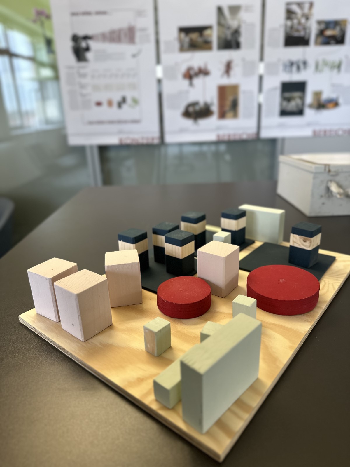 Ein hölzernes Architekturmodell mit rechteckigen und runden Blöcken steht auf einem Tisch; im Hintergrund hängen an einer Wand unscharfe Architekturplakate über Bürokonzepte der Zukunft. at Hochschule Coburg