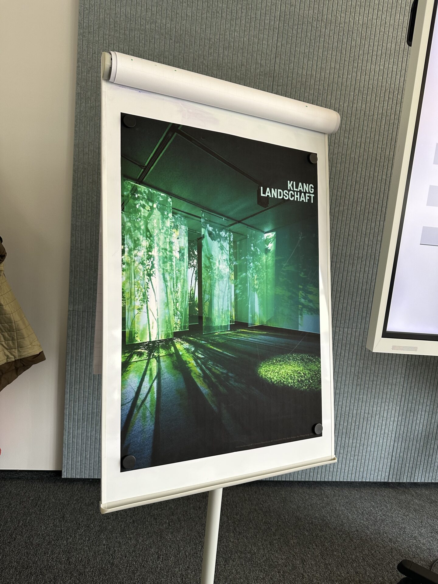 Ein Plakat auf einem Flipchartständer zeigt einen Raum mit grünen Lichtprojektionen, die an einen Wald erinnern - eine innovative Anspielung auf Bürokonzepte der Zukunft. Rechts oben steht "KLANG LANDSCHAFT", im Hintergrund eine gerippte graue Wand und ein Teil eines Whiteboards. at Hochschule Coburg