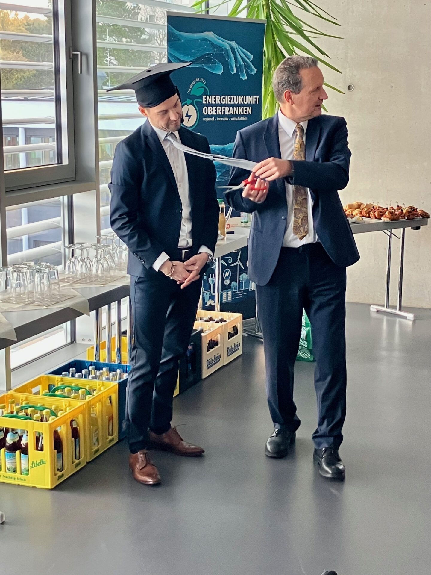 Zwei Männer in Anzügen stehen in einem Haus. Der eine trägt eine Abschlussmütze und lässt sich von dem anderen eine Schleife umbinden. Getränke in Kisten und Snacks stehen in der Nähe. Im Hintergrund steht ein Schild mit der Aufschrift "Energiezukunft Oberfranken" und signalisiert damit die Ausrichtung auf die Zukunft. at Hochschule Coburg