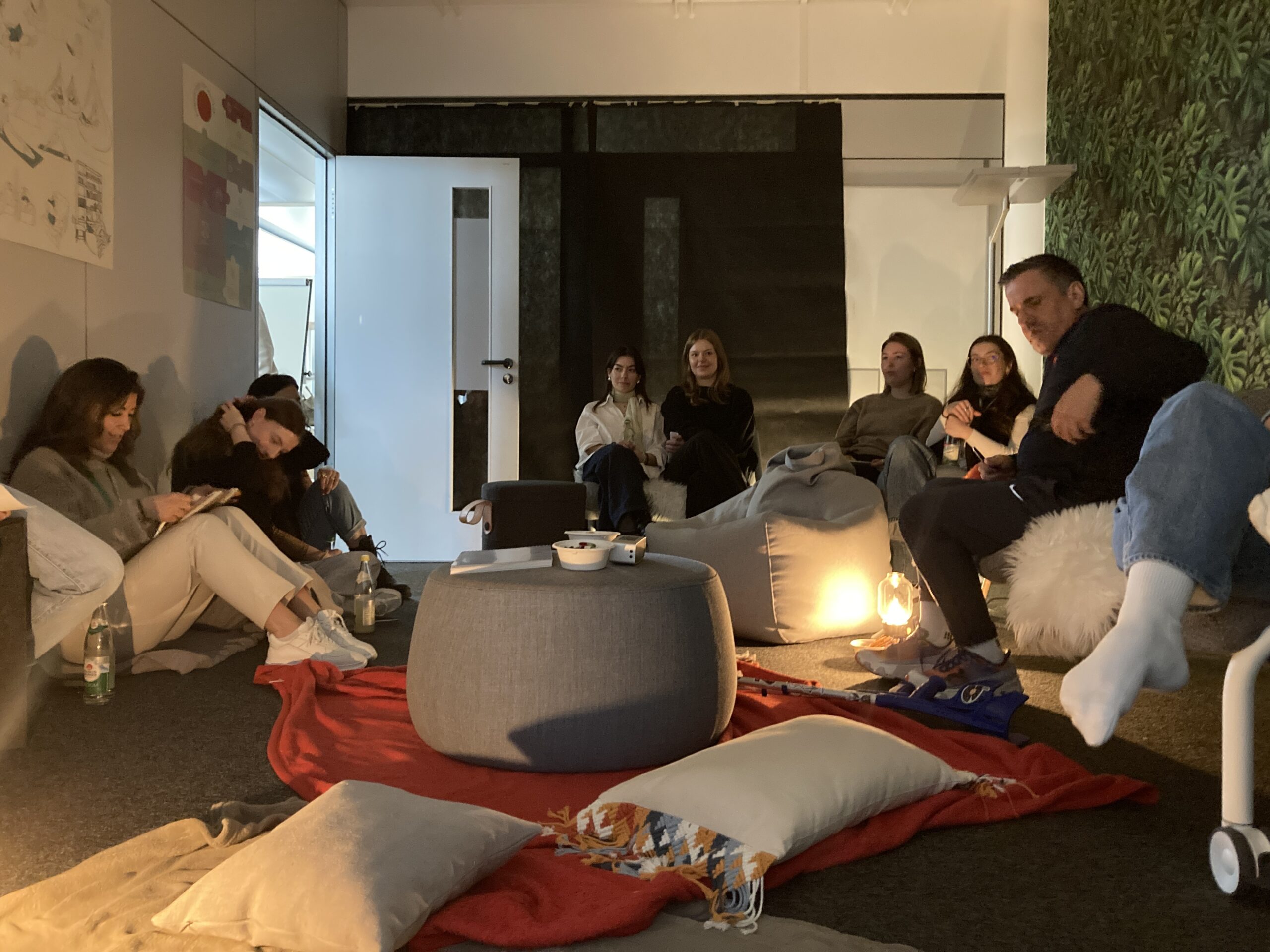 In Anlehnung an die Bürokonzepte der Zukunft entspannen sich die Menschen auf Sitzsäcken und Kissen in einem gemütlichen, schwach beleuchteten Raum mit Snacks und einer grünen, begrünten Wand - einige plaudern, andere checken ihre Telefone, was verdeutlicht, dass Arbeitsräume permanent im Wandel sind. at Hochschule Coburg