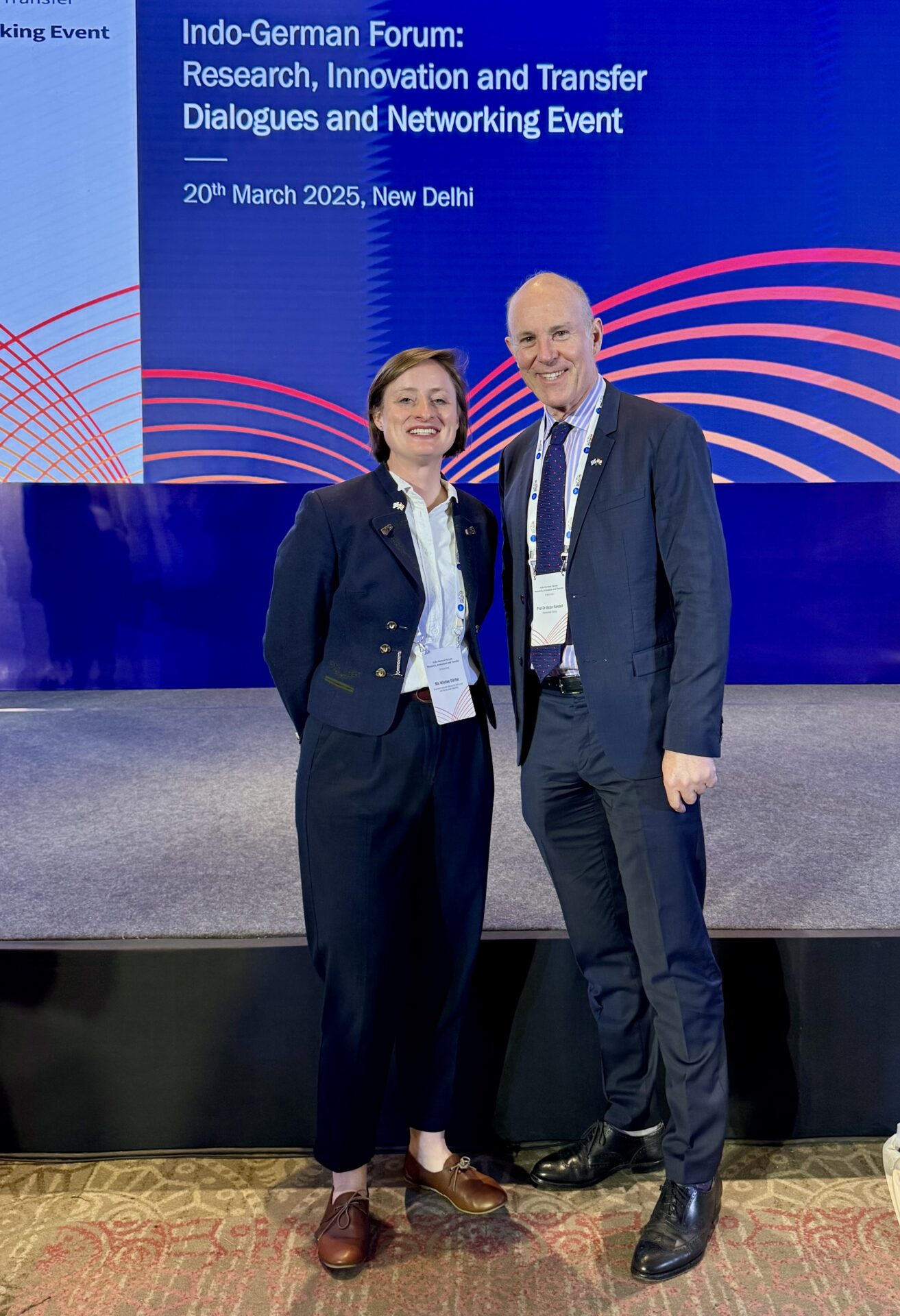 Zwei Personen in Business-Kleidung stehen lächelnd zusammen vor einem Bühnenhintergrund mit Text über das Indo-German Forum: Research, Innovation and Transfer Dialogues, showcasing international Präsenz am 20. März 2025 in Neu Delhi. at Hochschule Coburg
