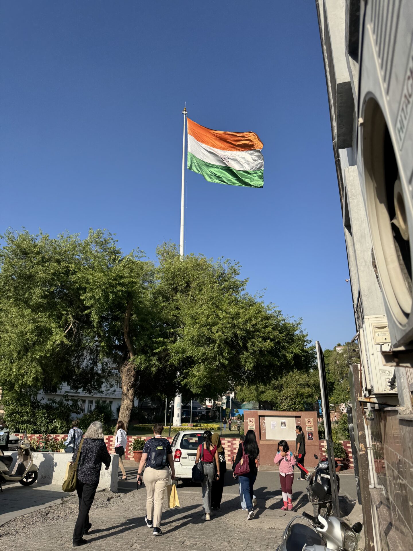 Eine Gruppe von Menschen geht im Freien in der Nähe von geparkten Fahrzeugen und einem großen Baum spazieren. Ein hoher Fahnenmast mit der indischen Nationalflagge weht vor einem strahlend blauen Himmel und symbolisiert die internationale Präsenz und die Möglichkeit zum Doppel-MBA-Networking. at Hochschule Coburg