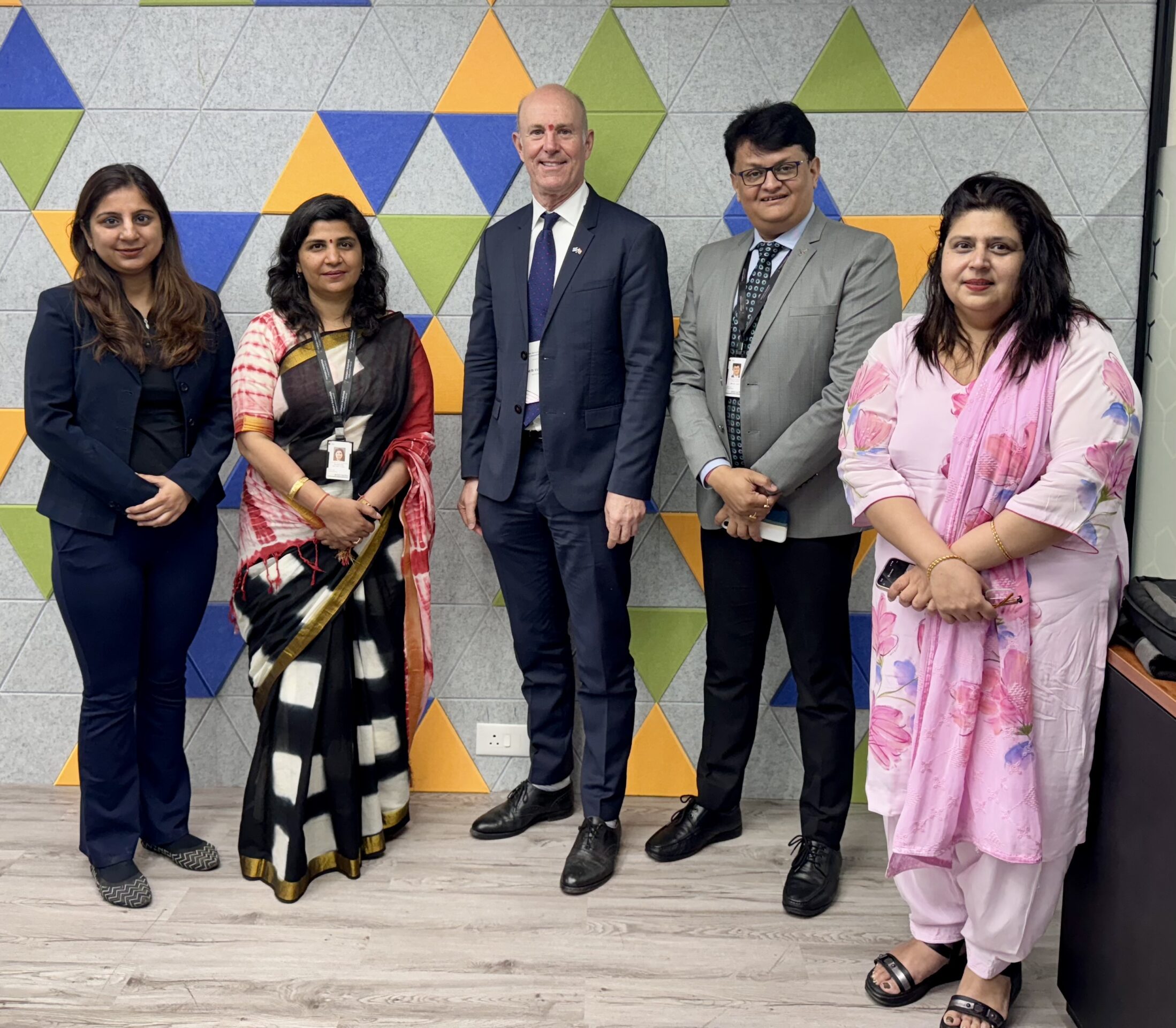 Fünf Personen, drei Frauen und zwei Männer, stehen nebeneinander in Business-Kleidung und lächeln in die Kamera. Im Hintergrund ist eine dekorative Wand mit blauen, grünen und gelben geometrischen Mustern zu sehen, die ihre internationale Präsenz widerspiegeln. at Hochschule Coburg