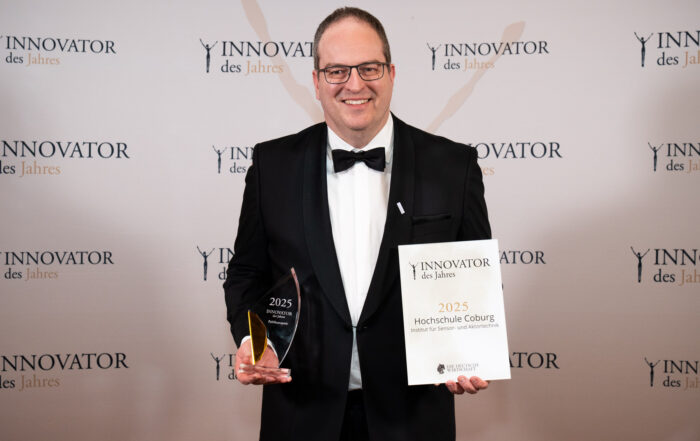 Ein Mann im Smoking lächelt, während er eine gläserne Auszeichnung und eine Urkunde mit der Aufschrift "Innovator des Jahres 2025 Hochschule Coburg" hält. Im Hintergrund sind wiederholte "Innovator des Jahres"-Logos zu sehen. at Hochschule Coburg