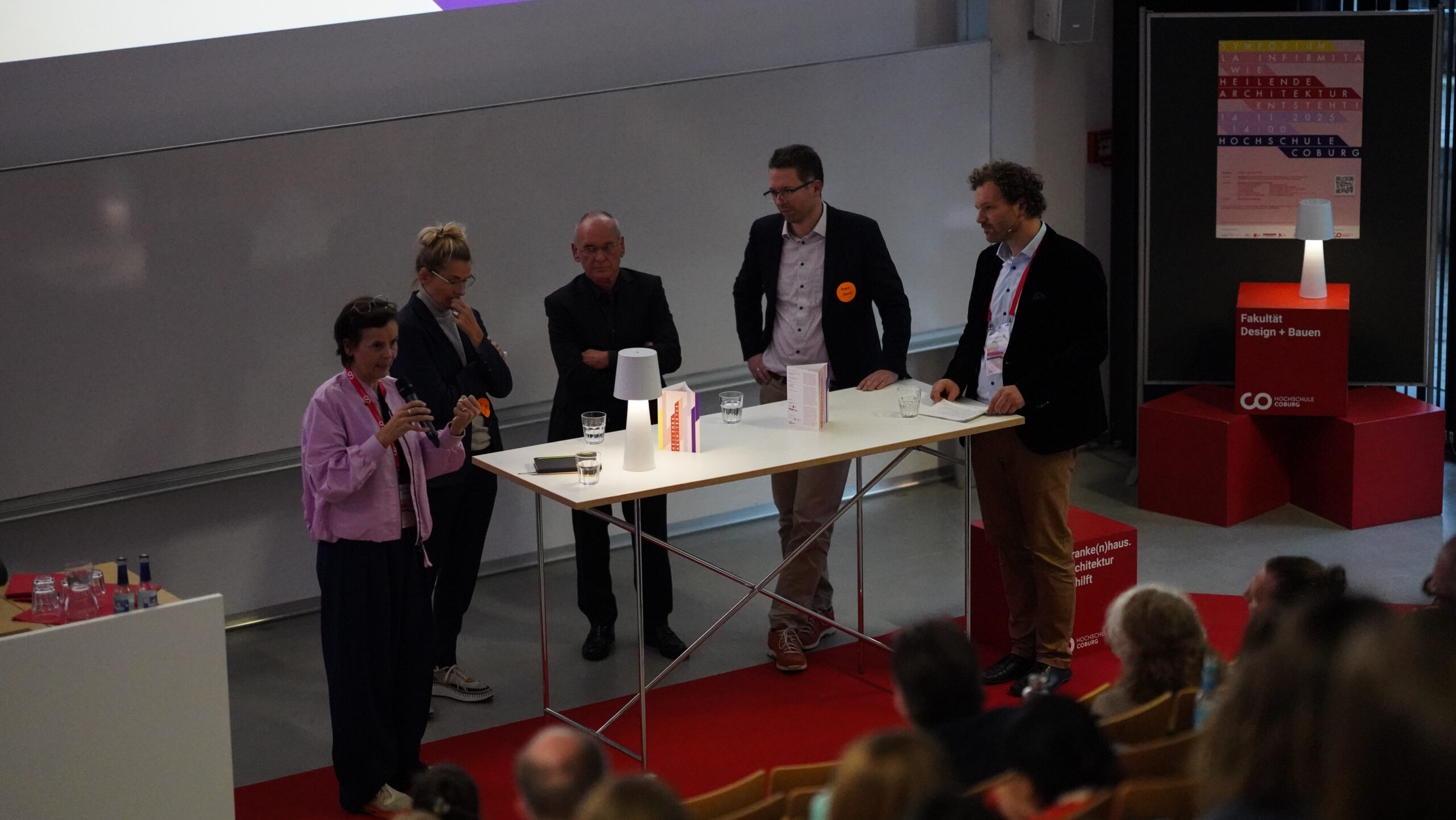 Bei einem Symposium über Krankenhausarchitektur stehen fünf Personen um einen hohen weißen Tisch auf der Bühne und unterhalten sich mit dem Publikum. Eine Frau hält ein Mikrofon, während die anderen zuhören. Auf dem Tisch liegen Papiere, eine kleine Lampe und ein Schild. at Hochschule Coburg