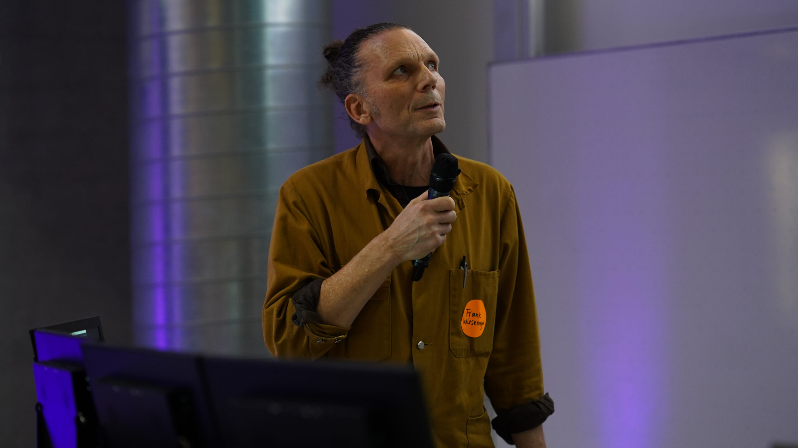 Eine Person mit langem zurückgebundenem Haar, brauner Jacke und Mikrofon in der Hand steht vor einer Tafel und einer großen Metallsäule und scheint auf einem modernen Symposium über Krankenhausarchitektur zu sprechen oder zu präsentieren. at Hochschule Coburg