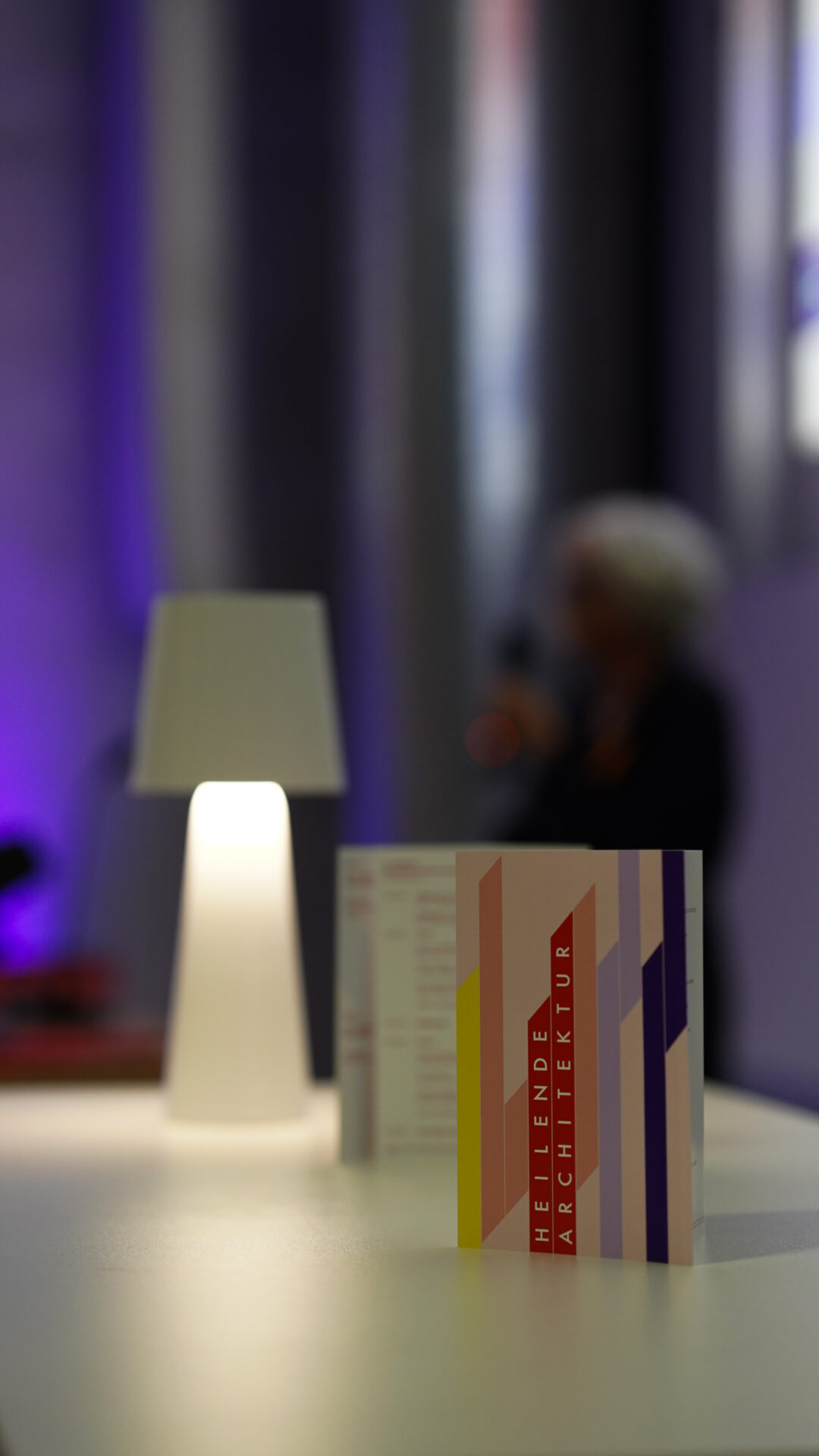 Eine Nahaufnahme einer Karte mit bunten vertikalen Streifen und dem Text "HEILENDE ARCHITEKTUR" auf einem weißen Tisch neben einer modernen Lampe, die in violettes Licht getaucht ist - und den kreativen Impuls von Symposien über innovative Krankenhausarchitektur einfängt. at Hochschule Coburg