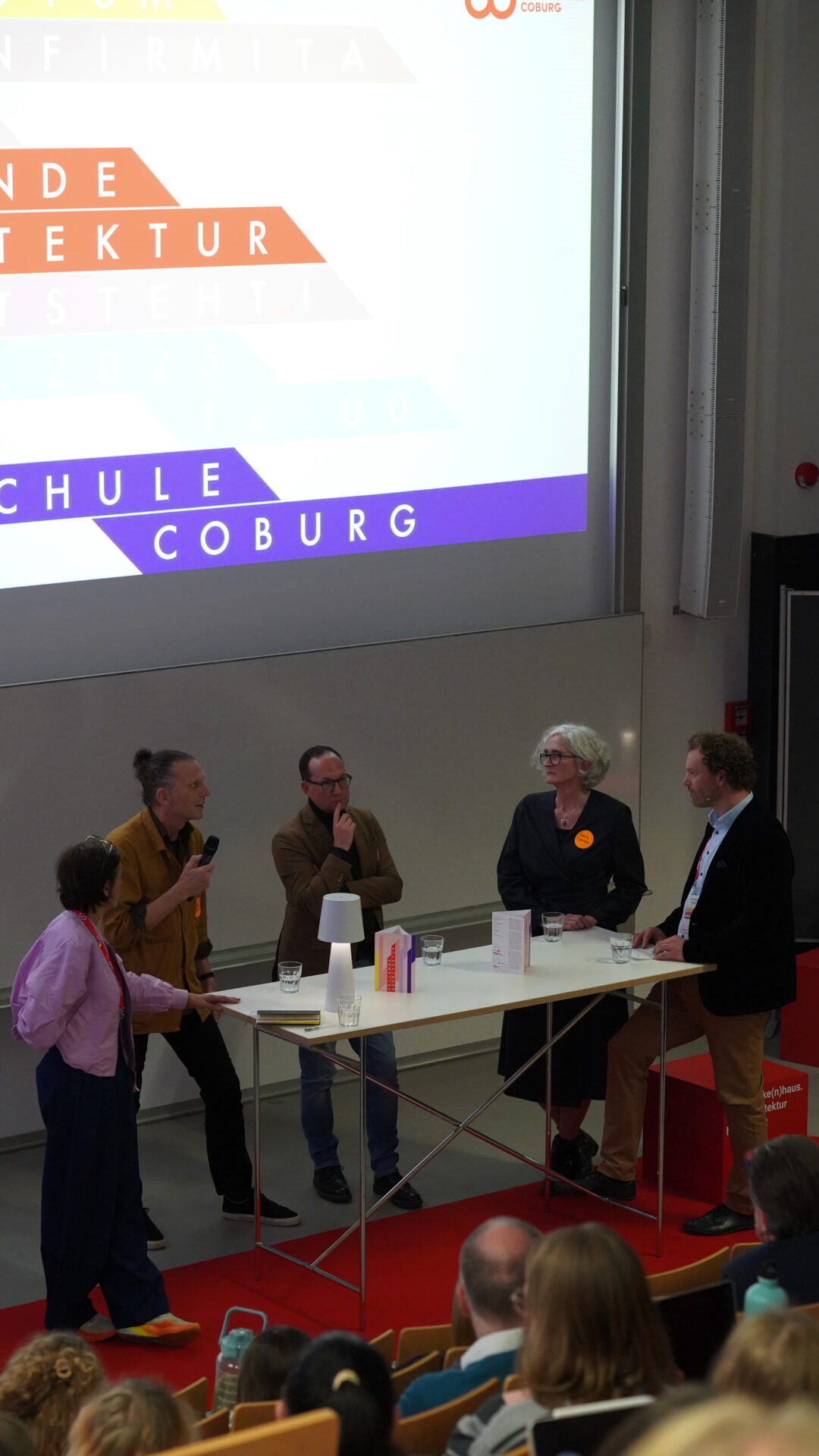 Fünf Personen führen eine Podiumsdiskussion während eines Krankenhausarchitektur-Symposiums vor einem Publikum in einem Hörsaal, das um einen Tisch mit Mikrofonen und Unterlagen herumsteht. Hinter ihnen ist eine große bunte Präsentationsfolie zu sehen. at Hochschule Coburg