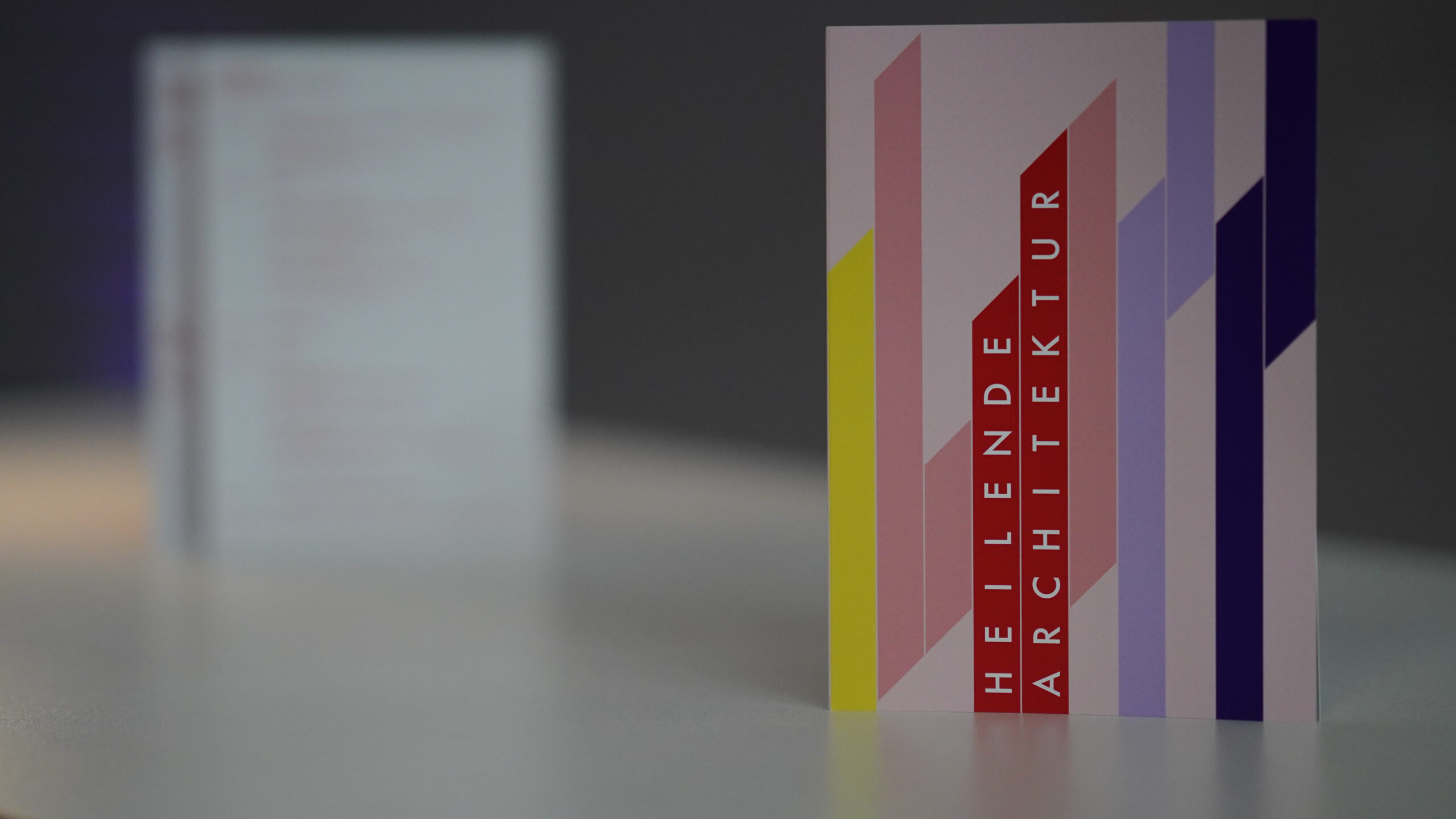 Eine farbenfrohe Karte mit geometrischen Mustern und vertikalen Streifen in Gelb, Rot, Rosa, Lavendel und Violett zeigt den Text "HEILENDE ARCHITEKTUR" auf einem roten Streifen und steht auf einer weißen Fläche - perfekt für ein Symposium über Krankenhausarchitektur. at Hochschule Coburg