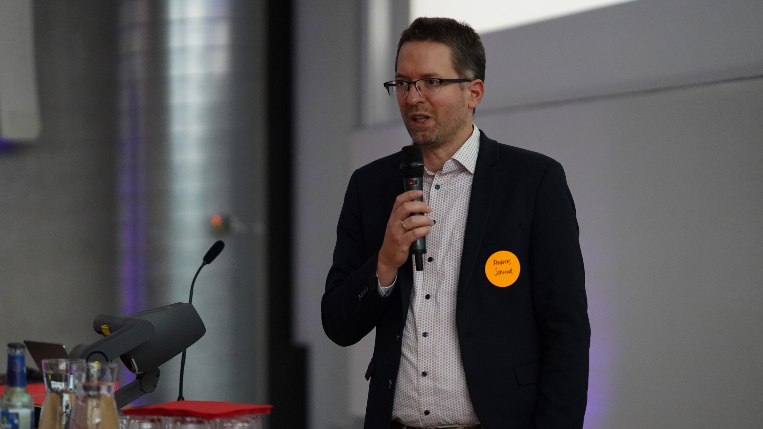 Ein Mann im Anzug und mit Brille hält ein Mikrofon, während er auf einem Symposium der Krankenhausarchitektur spricht. Er trägt ein gelbes Namensschild und steht an einem Podium mit Wasserflaschen und einem Mikrofon, wobei der Hintergrund unscharf ist. at Hochschule Coburg