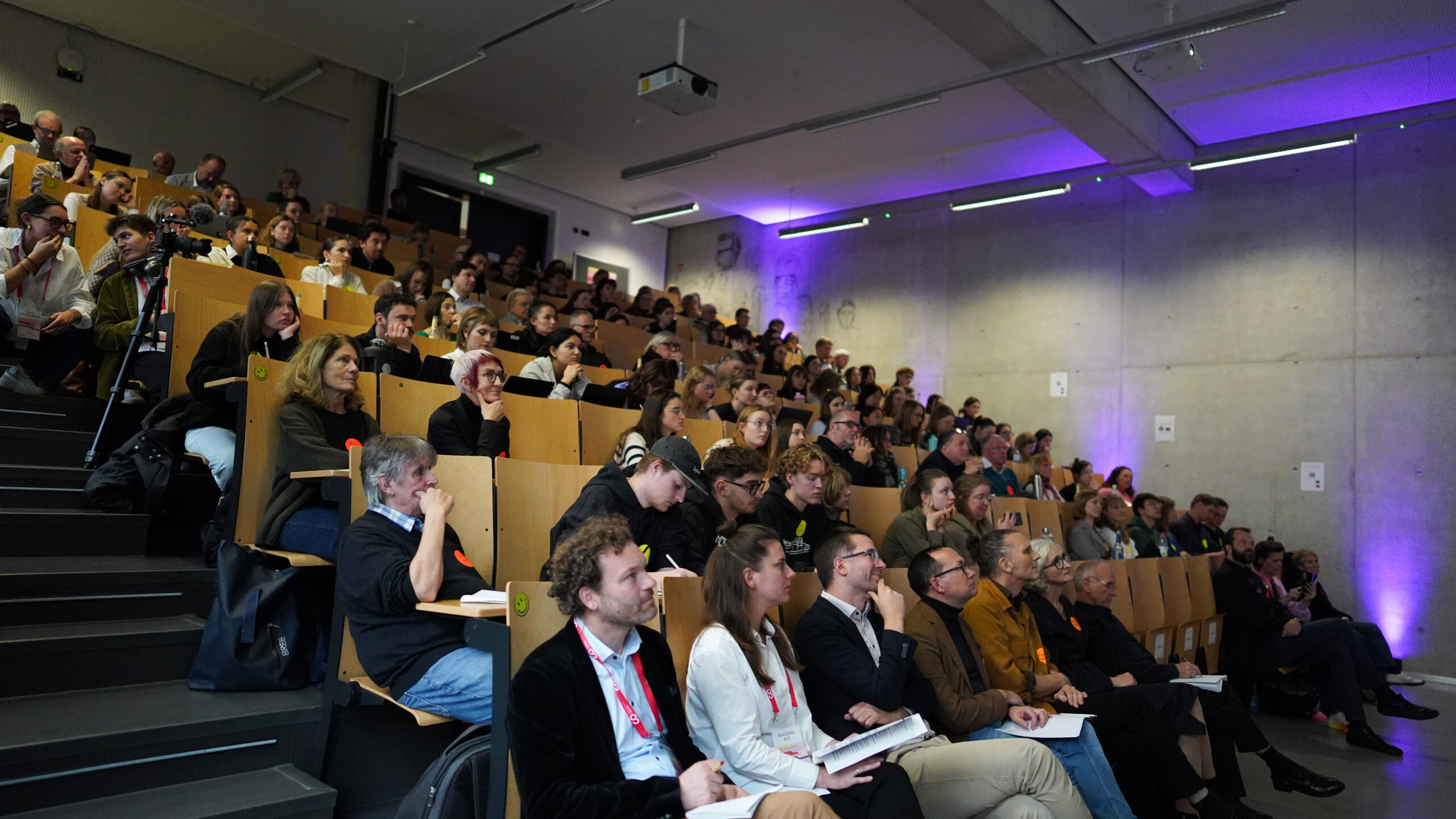 Eine große Gruppe von Menschen sitzt auf gestuften Holzstühlen und verfolgt aufmerksam ein Symposium über Krankenhausarchitektur in einem modernen Hörsaal mit Betonwänden und violetter Beleuchtung. at Hochschule Coburg