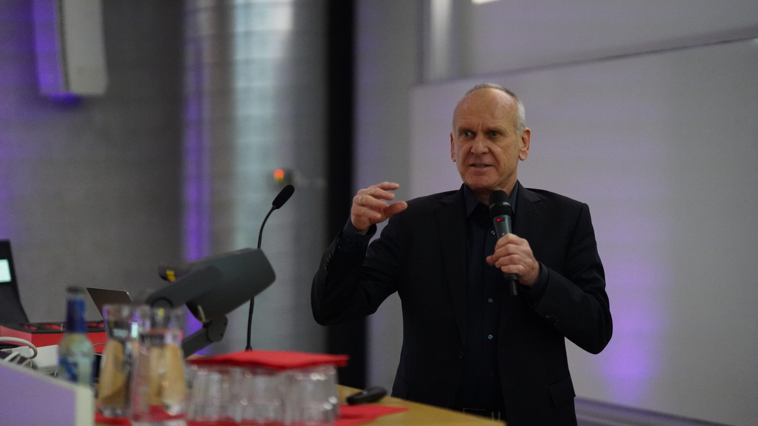 Auf einem Symposium der Krankenhausarchitektur spricht ein Mann in einem dunklen Anzug in ein Mikrofon, während er am Podium gestikuliert und dem Publikum frische Impulse gibt; im Vordergrund sind Gläser, Flaschen und rote Ordner zu sehen. at Hochschule Coburg