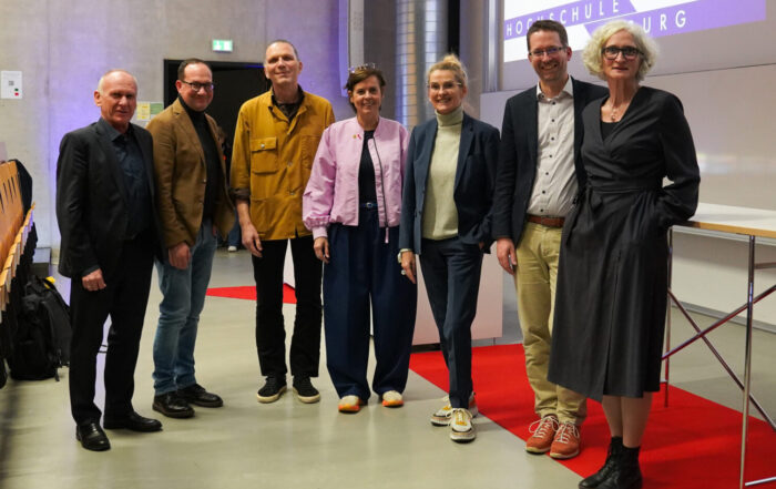Sieben Personen stehen in einer Reihe auf einem roten Teppich in einem modernen Hörsaal und posieren für ein Gruppenfoto vor den Präsentationsbildschirmen nach einem Symposium der Krankenhausarchitektur, mit leeren Holzstühlen im Hintergrund. at Hochschule Coburg