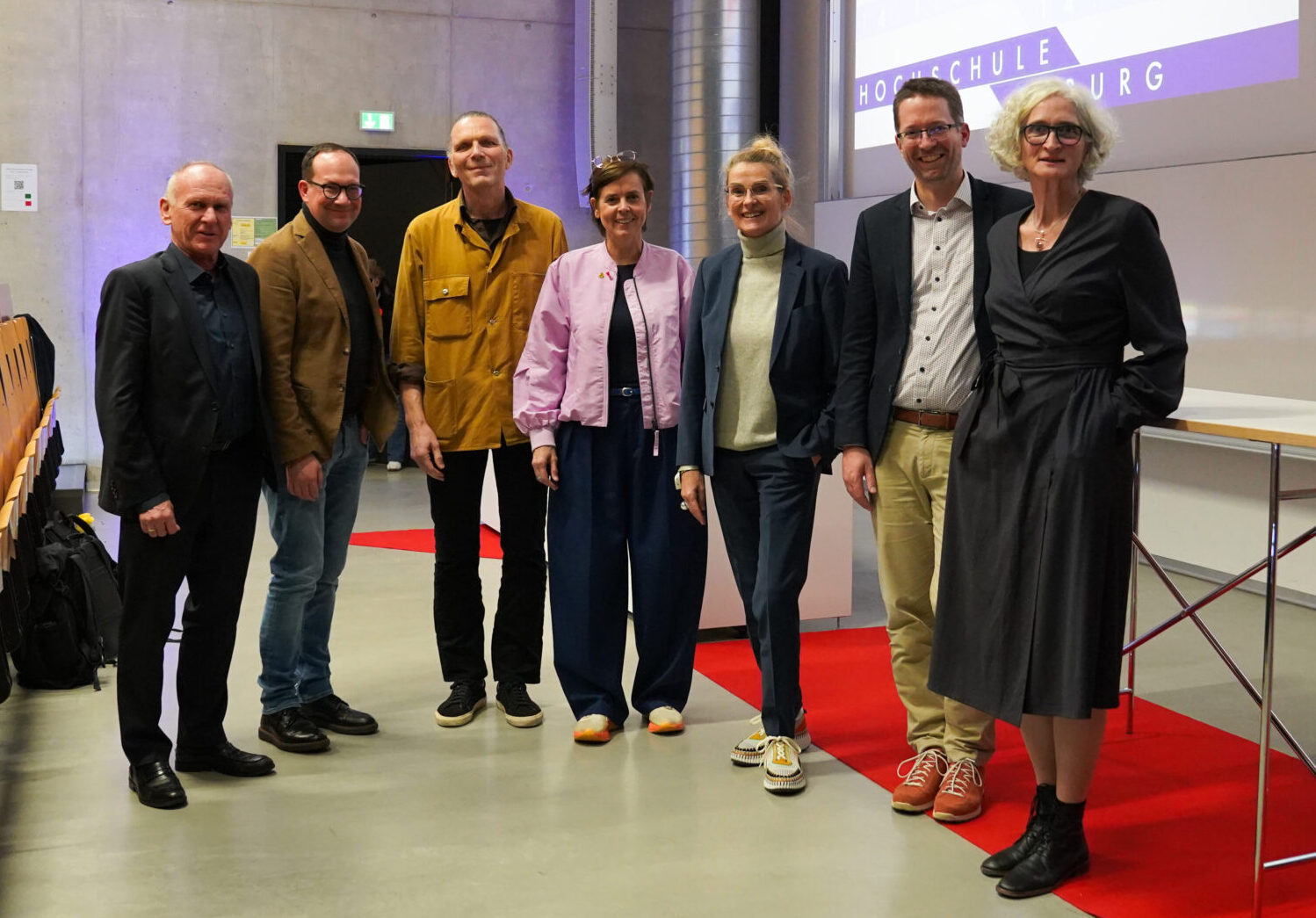 Sieben Personen stehen in einer Reihe auf einem roten Teppich in einem modernen Hörsaal und posieren für ein Gruppenfoto vor den Präsentationsbildschirmen nach einem Symposium der Krankenhausarchitektur, mit leeren Holzstühlen im Hintergrund. at Hochschule Coburg