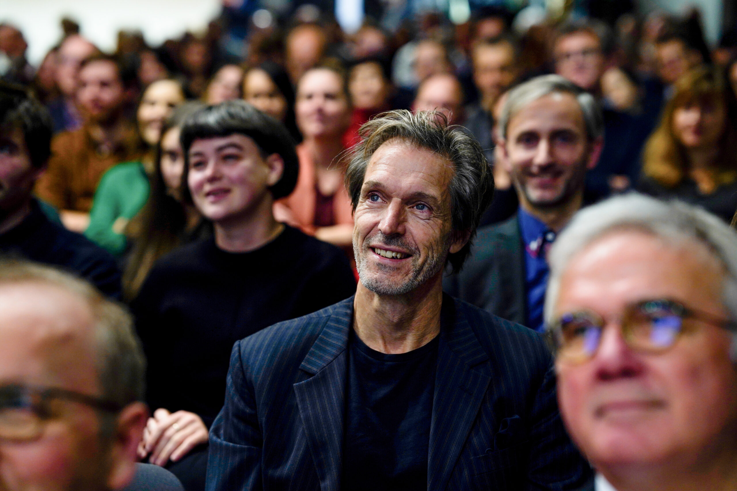 Ein lächelnder Mann in einem dunklen Anzug sitzt inmitten eines bunt gemischten, aufmerksamen Publikums bei einer Indoor-Veranstaltung der Hochschule, möglicherweise Dies academicus 2025, mit verschwommenen Gesichtern der um ihn herum sitzenden Personen. at Hochschule Coburg