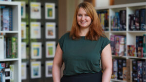 A woman with long light brown hair wearing a dark green shirt stands smiling in a library or bookstore, with shelves of books and board games in the background. at Hochschule Coburg