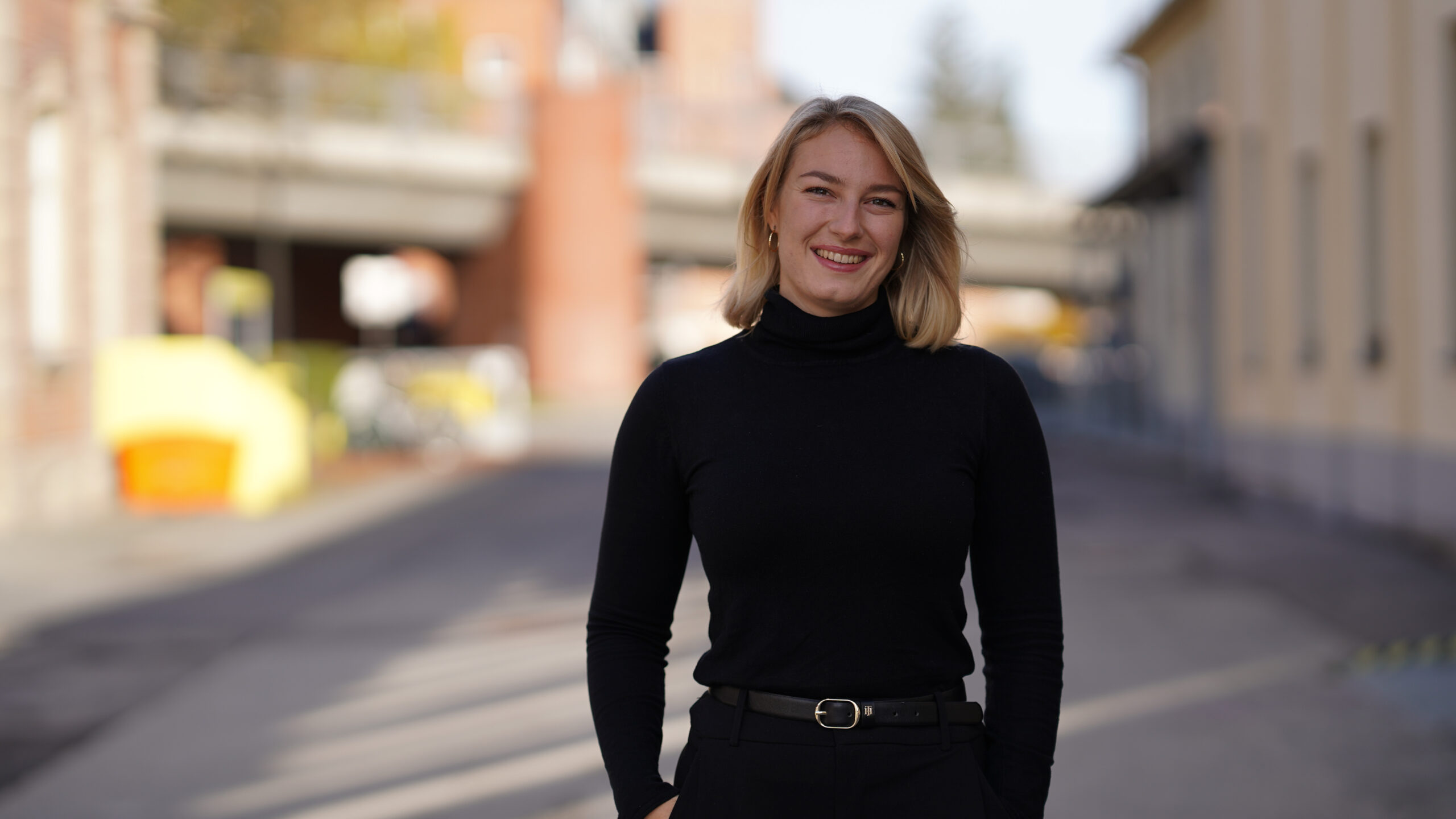 Eine Person mit blondem Haar, die einen schwarzen Rollkragenpullover und eine schwarze Hose trägt, steht lächelnd in einer städtischen Umgebung im Freien mit unscharfen Gebäuden und einer Straße im Hintergrund. at Hochschule Coburg
