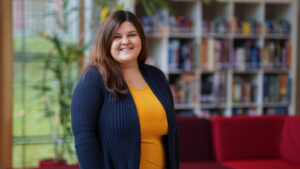 A woman with long brown hair, wearing a yellow top and navy blue cardigan, stands smiling in a brightly lit room with bookshelves and red couches in the background. at Hochschule Coburg