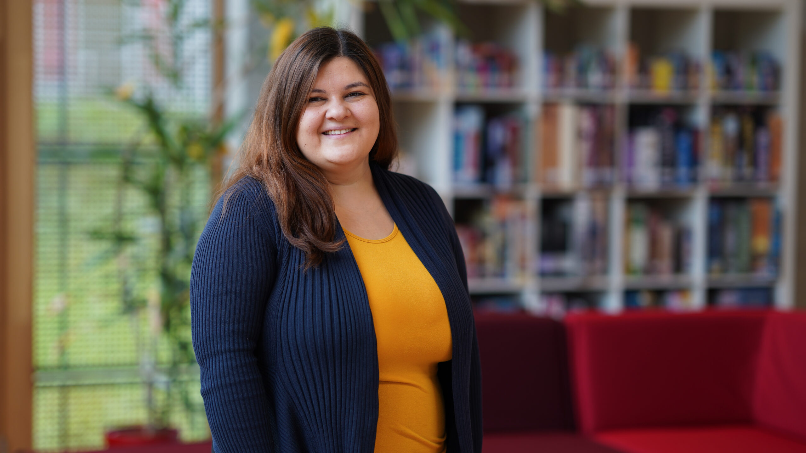 Eine Frau mit langen braunen Haaren, die ein gelbes Oberteil und eine marineblaue Strickjacke trägt, steht lächelnd in einem Innenraum vor Bücherregalen und roten Sofas. at Hochschule Coburg