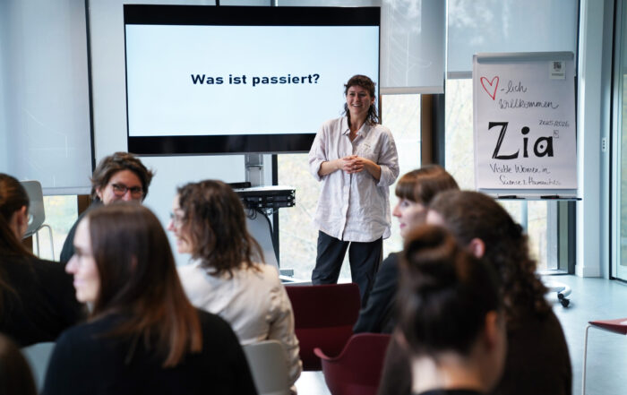 Eine Frau steht lächelnd vor einem sitzenden Publikum in einem modernen Raum auf dem ZIA-Kongress 2025. Auf einem Bildschirm hinter ihr steht "Was ist passiert?" auf Deutsch, während auf einem Flipchart neben ihr handschriftliche Notizen zu lesen sind, darunter das Wort "Zia". at Hochschule Coburg