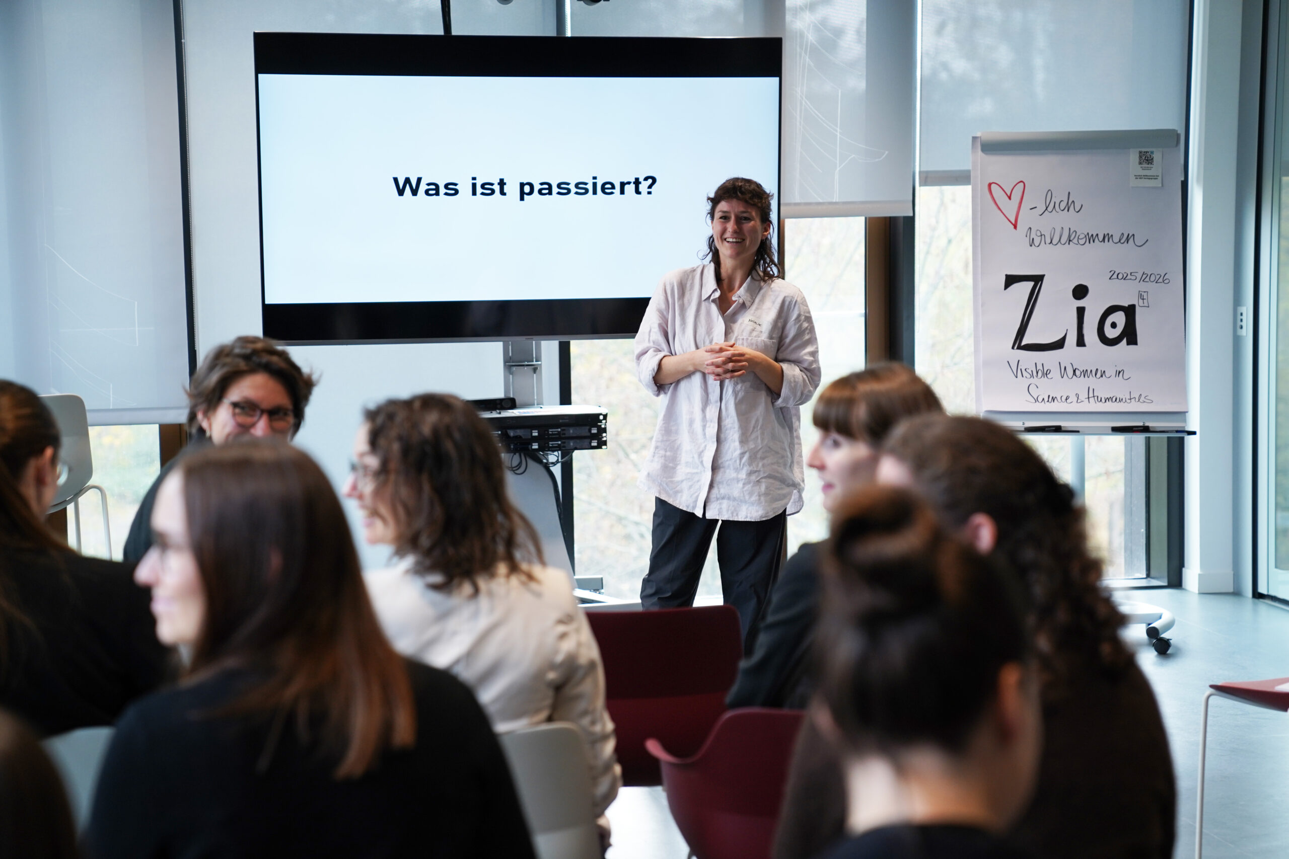 Eine Frau steht lächelnd vor einem sitzenden Publikum in einem modernen Raum auf dem ZIA-Kongress 2025. Auf einem Bildschirm hinter ihr steht "Was ist passiert?" auf Deutsch, während auf einem Flipchart neben ihr handschriftliche Notizen zu lesen sind, darunter das Wort "Zia". at Hochschule Coburg