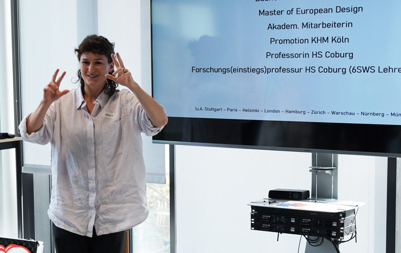 Eine Person steht vor einem Präsentationsbildschirm, auf dem akademische Titel in deutscher Sprache auf dem ZIA-Kongress 2025 angezeigt werden, und gestikuliert mit erhobenen Händen. Der Raum hat große Fenster mit Blick auf Bäume und moderne Büromöbel in Berlin. at Hochschule Coburg